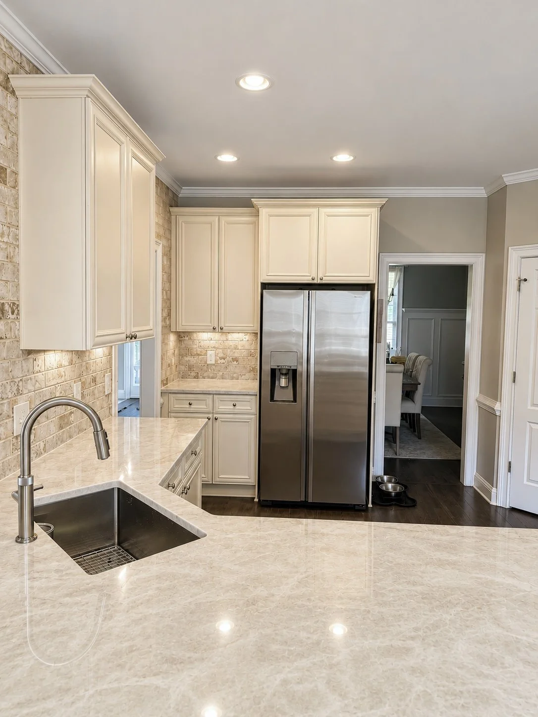 Kitchen with cream-colored cabinets, beige marble countertops, stainless steel refrigerator, black dog bowls on dark hardwood floor, exposed brick wall, recessed ceiling lights, view into dining room.