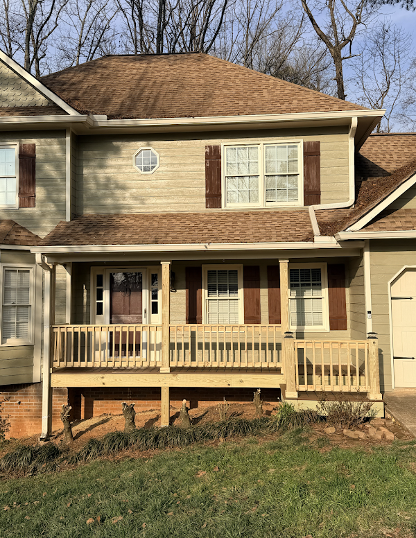 Front view of a two-story house with a new wooden front porch, beige siding, brown shutters, and a gabled roof, with leafless trees in the background.