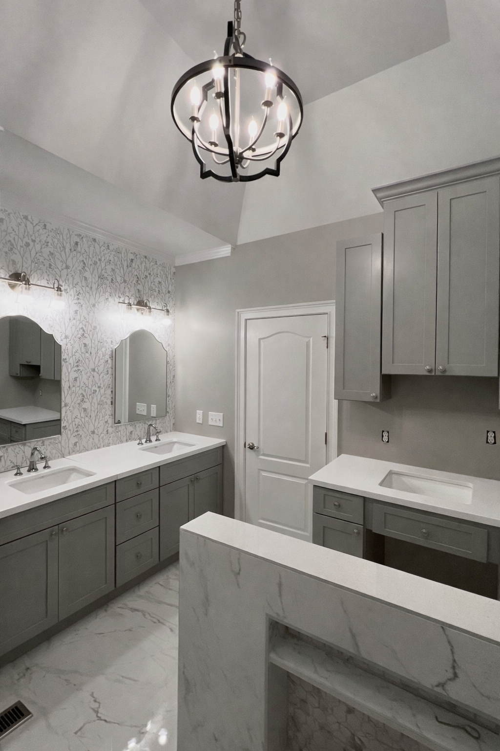 A modern bathroom featuring a black chandelier, double vanity sinks with mirrors, gray cabinetry, marble countertops, and a patterned gray wallpaper.