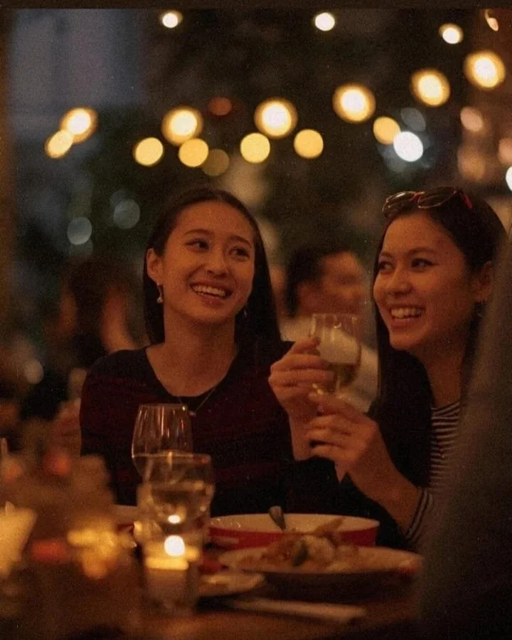 Two women smiling and toasting with drinks at a dimly lit restaurant or bar, with blurred lights and other people in the background.