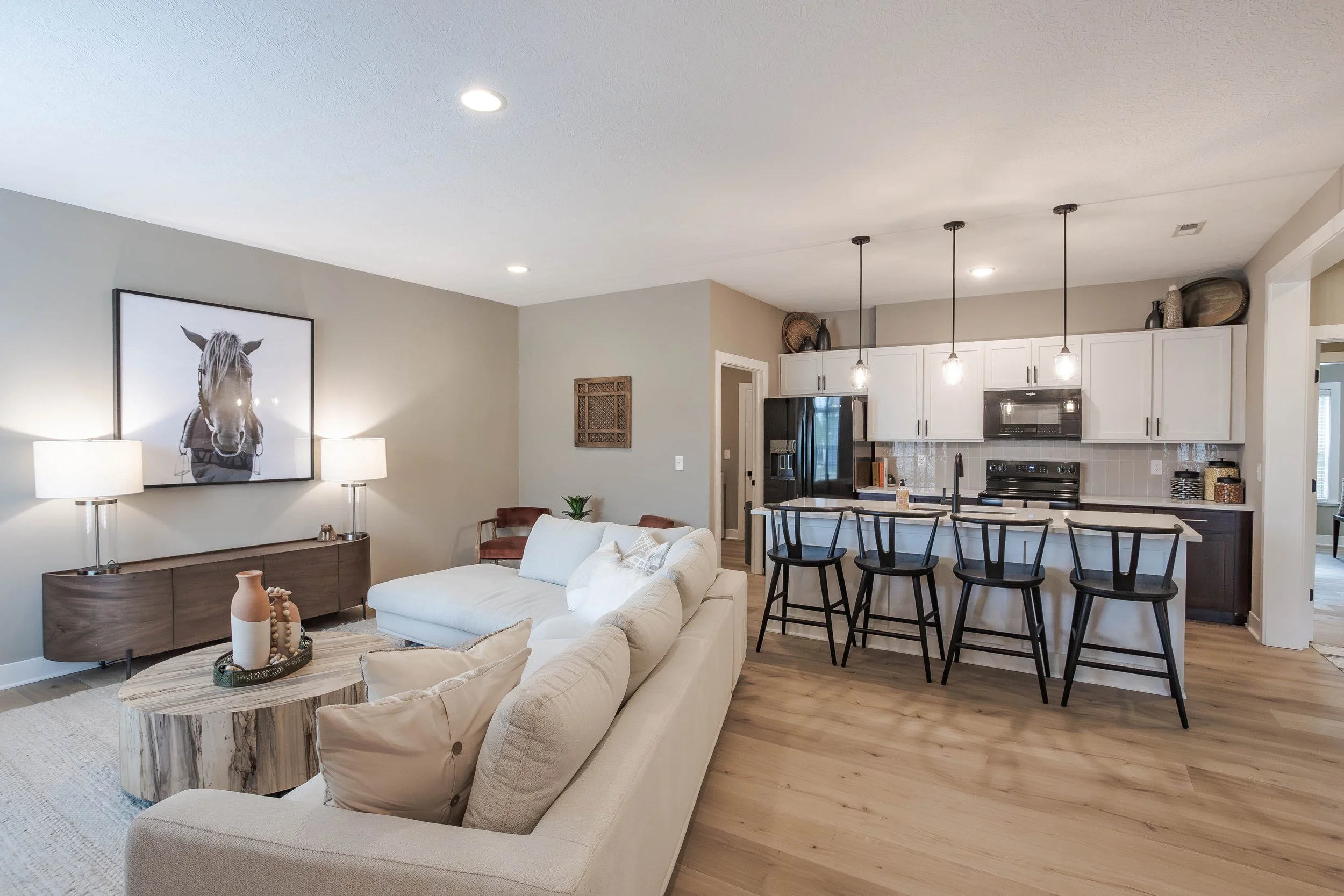 Open concept living room and kitchen with white cabinets, black appliances, and a breakfast bar with black chairs. Living area has a beige sofa, a wood coffee table, a console with lamps, and wall art featuring a horse.