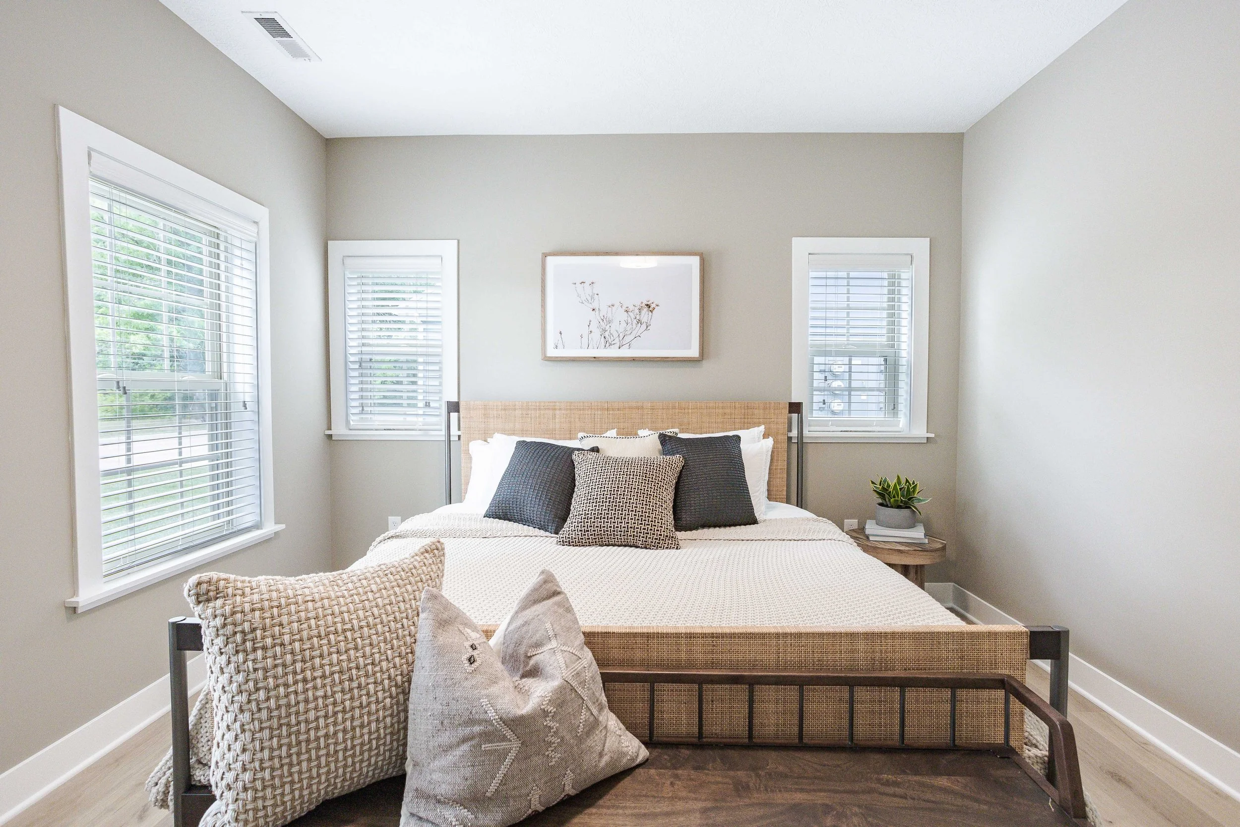A neatly made bed with a woven headboard, accented with multiple gray and beige pillows, in a bedroom with three windows and a framed art piece above the bed; a small nightstand with a potted plant and books, light-colored walls, and a window with blinds on each side of the bed.