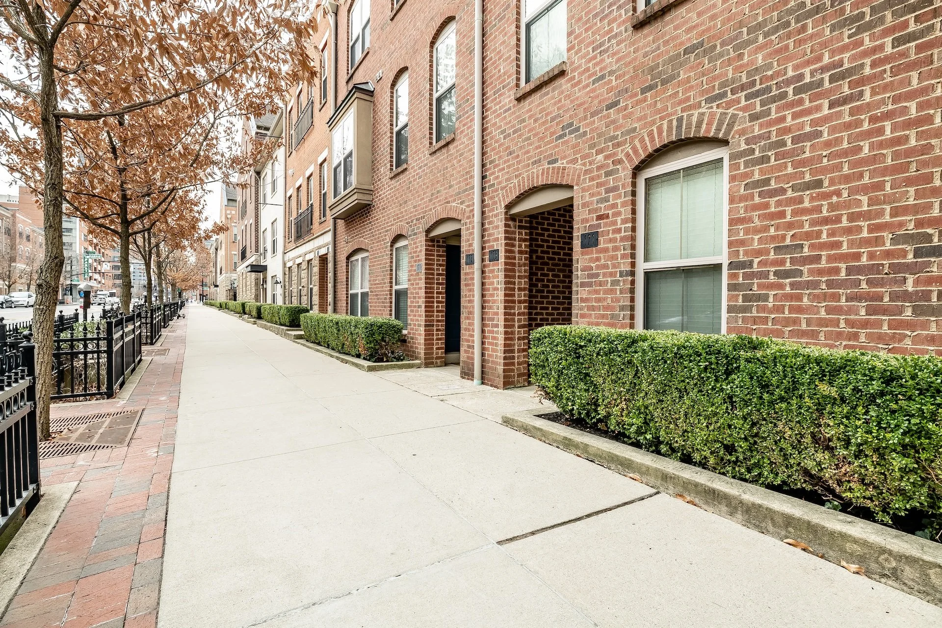 A downtown Columbus sidewalk lined with green bushes, trees with brown leaves, and red brick buildings with windows and small balconies.