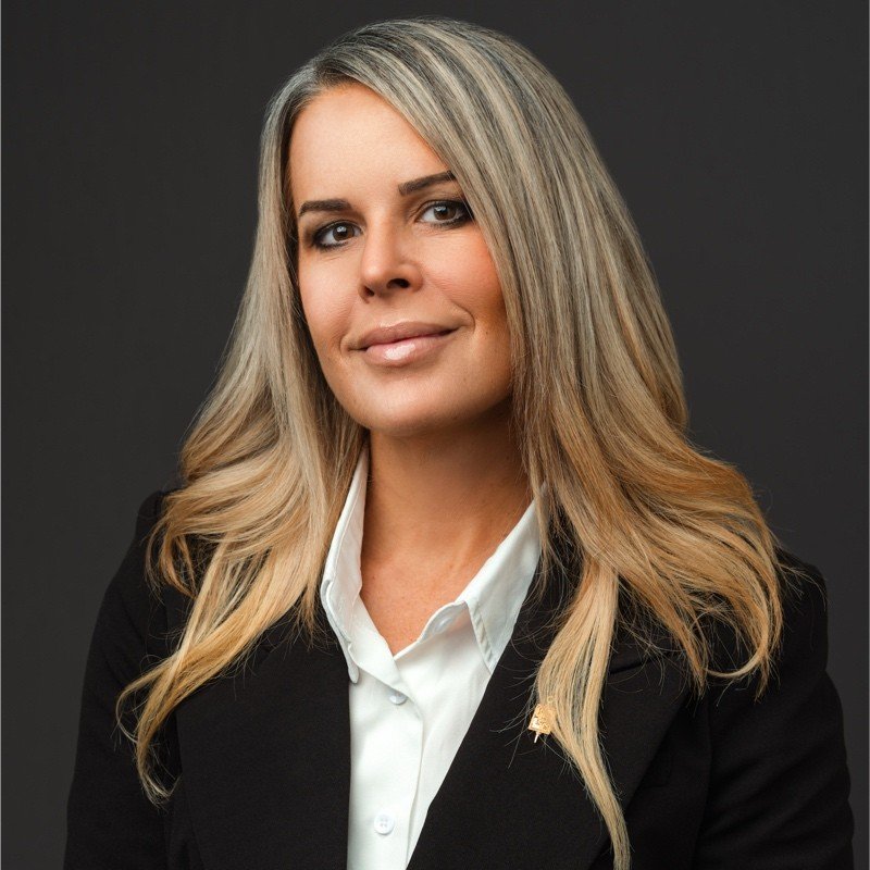 Professional woman with long, blonde hair, wearing a black blazer and white shirt, posing against a dark background.
