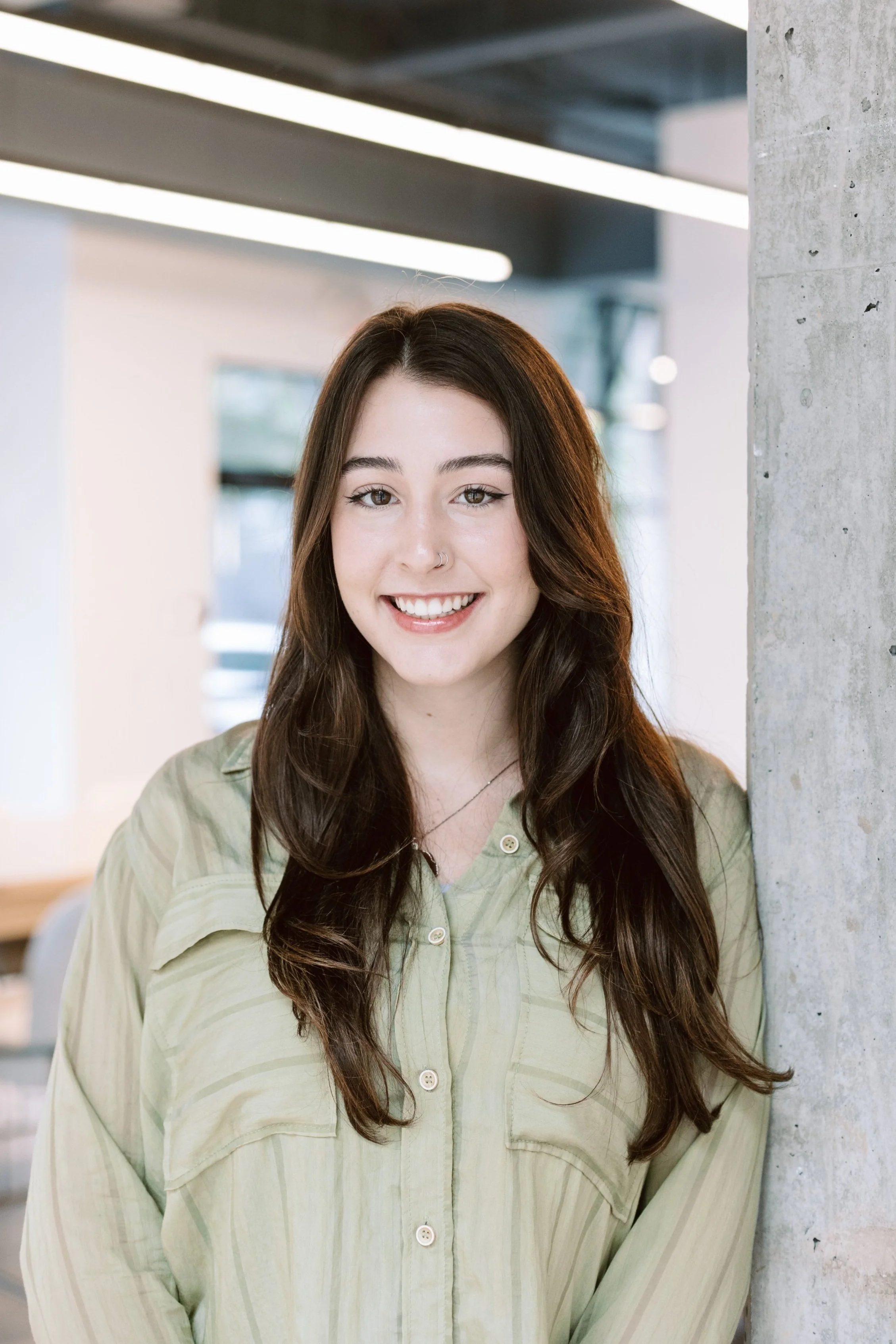 A young woman with long brown hair, wearing a light green button-up shirt, smiling and leaning against a concrete pillar indoors.