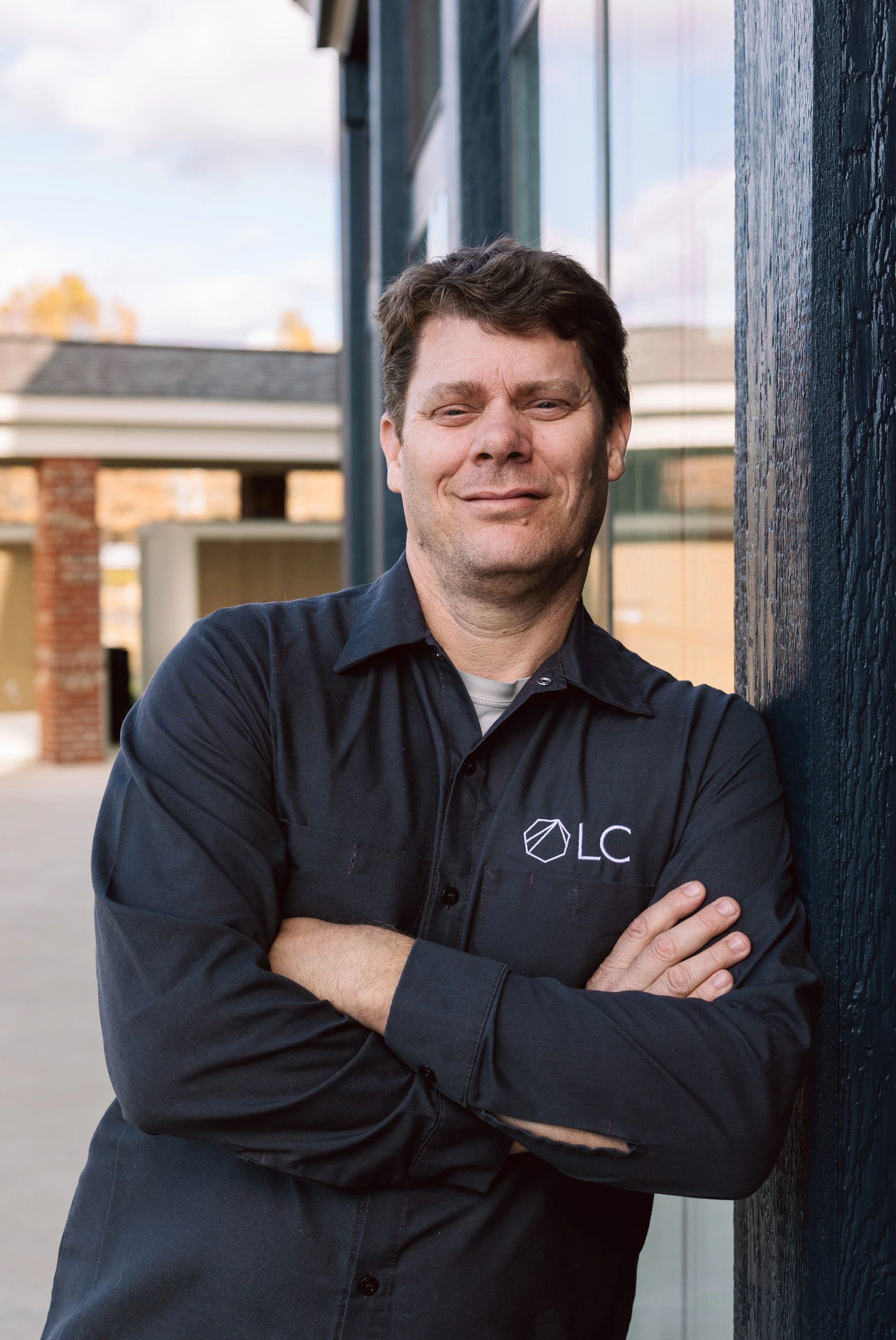 A man in a dark shirt with a logo and initials 'OLC' is leaning against a dark blue wall outdoors, with arms crossed, smiling confidently.
