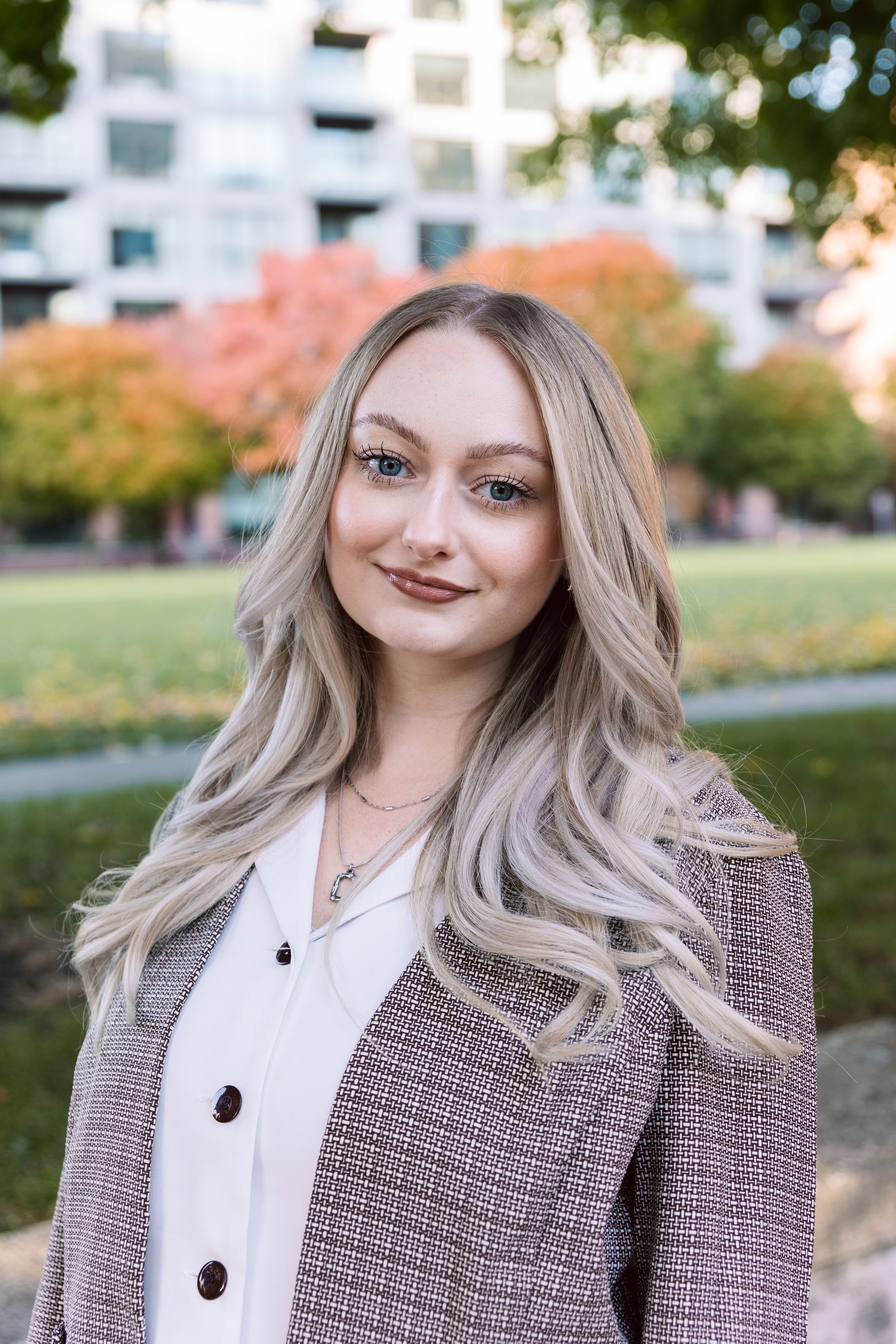 A young woman with long blonde hair, blue eyes, and light makeup, smiling outdoors in front of colorful autumn trees and modern apartment buildings.