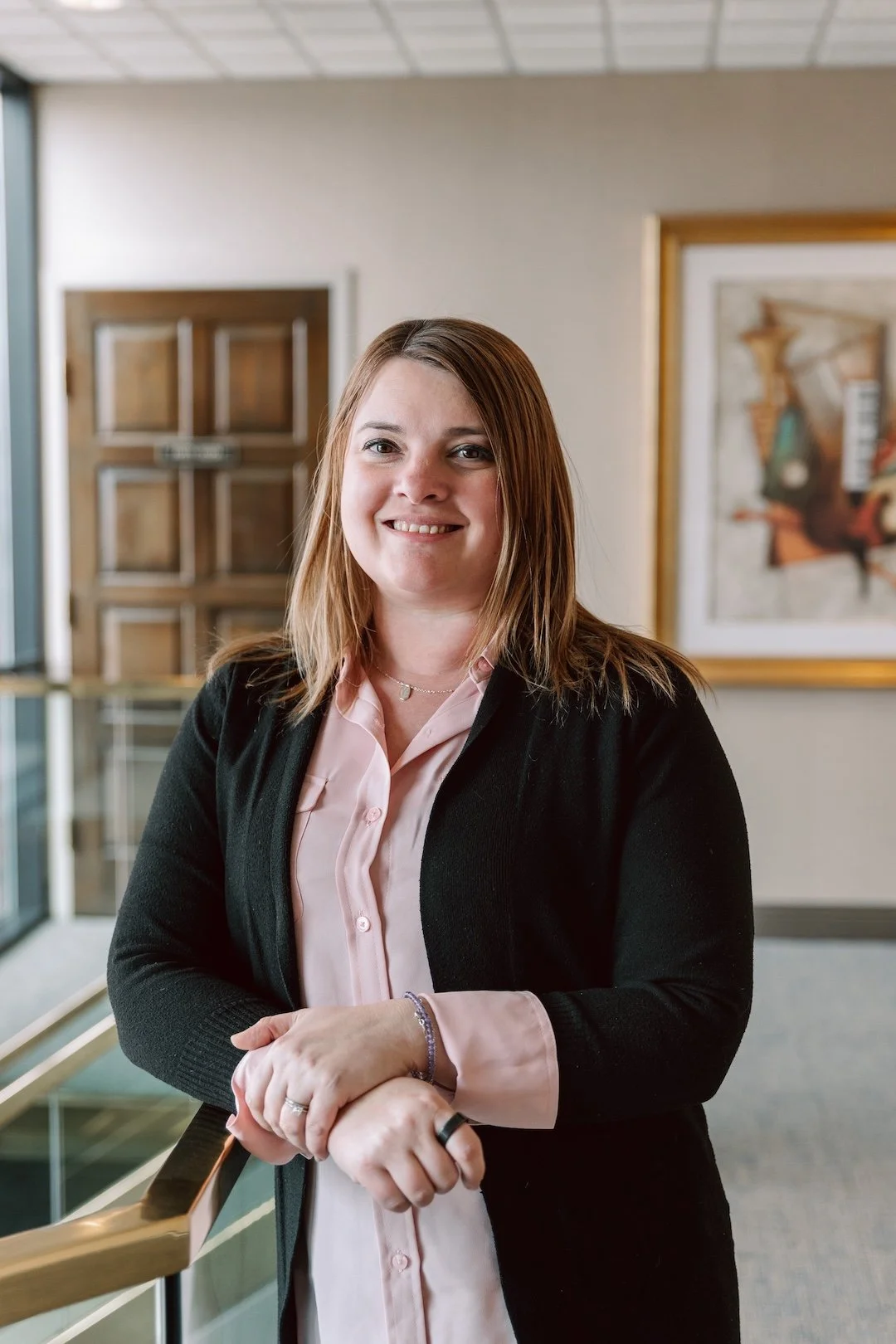 A woman with medium-length brown hair smiling, wearing a light pink collared shirt and a black cardigan, standing inside a building near a railing with artwork and wooden door in the background.