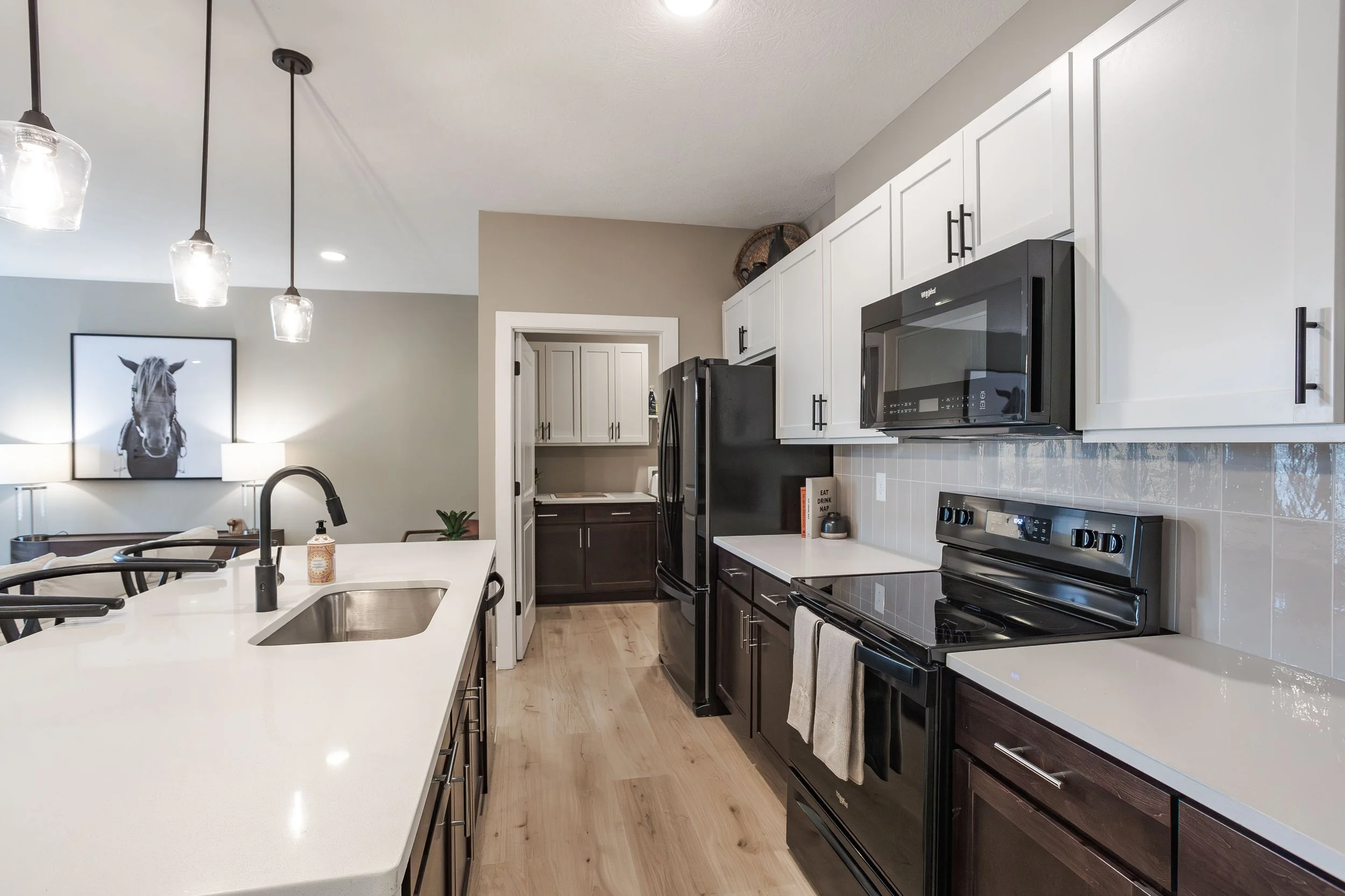 Modern kitchen with white upper cabinets and black lower cabinets, black appliances including a microwave, stove, and refrigerator, a white countertop with a sink, and hardwood floors. In the background, a dining area with a picture of a horse and a 
