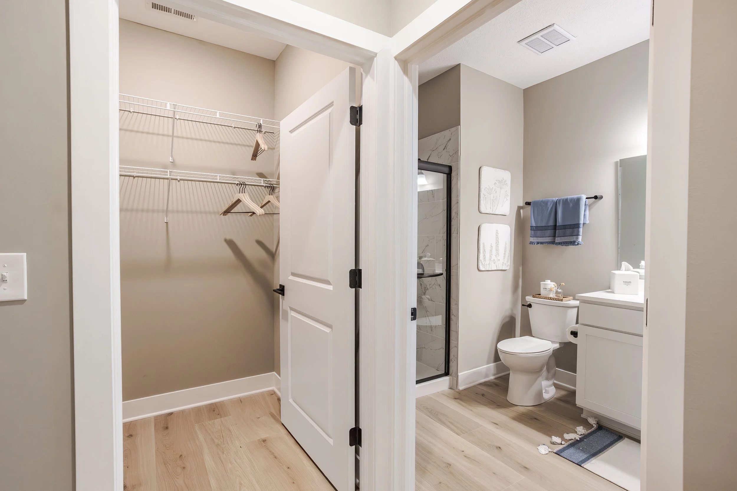 Bathroom with a toilet, white vanity, mirror, and decorations, and an adjacent empty walk-in closet with beige walls and wooden flooring.
