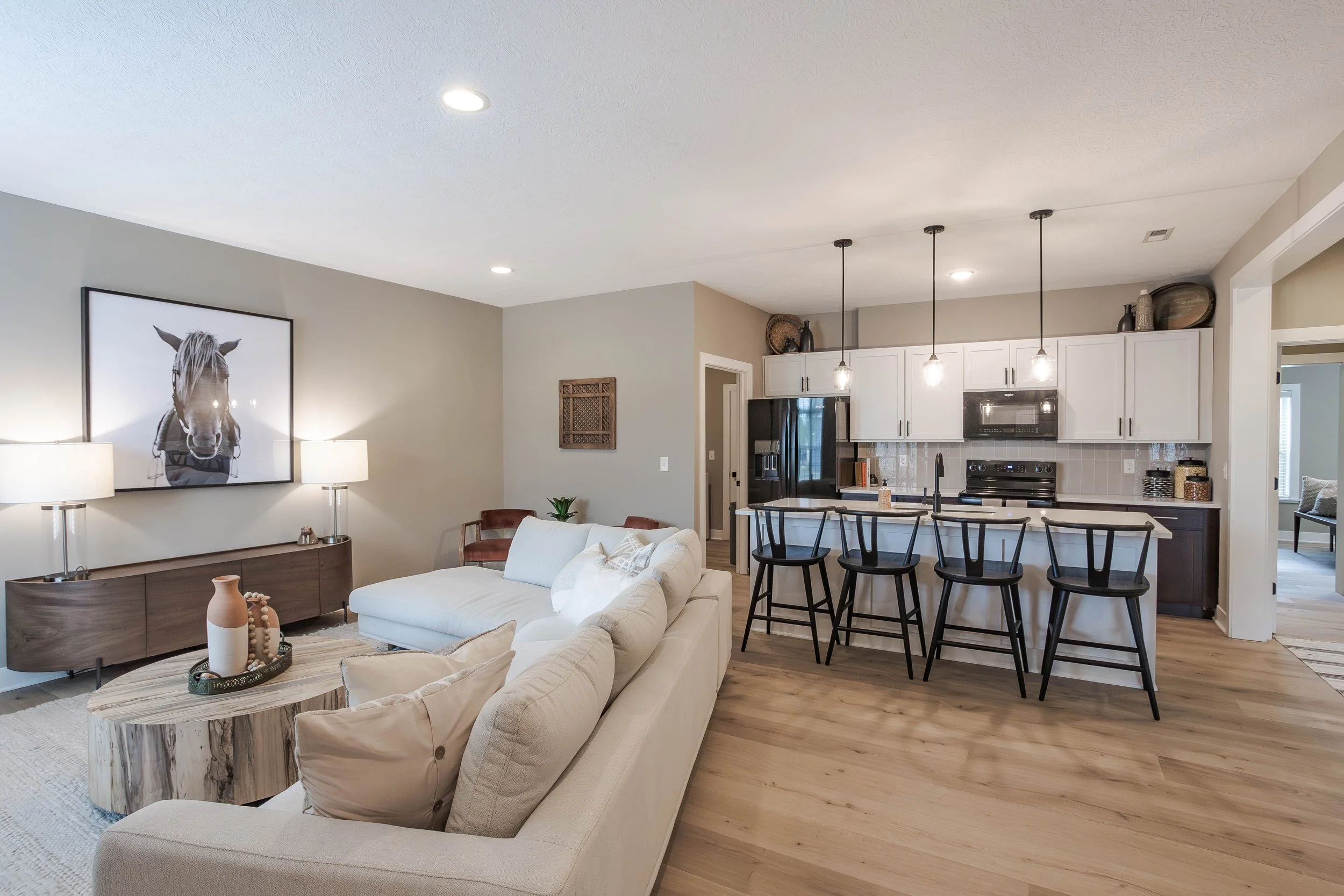 Open-concept living room and kitchen with white sofa, wooden coffee table, black bar stools, and pendant lights.