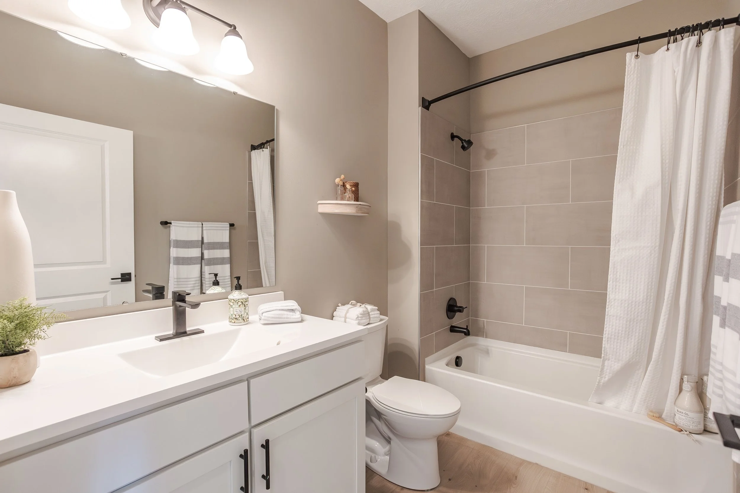 A modern bathroom with a white vanity, a large mirror, a single-handle faucet, and decorative items including soap dispenser and towels. There is a toilet next to the vanity and a bathtub with a beige tiled shower surround and a white curtain. The ro