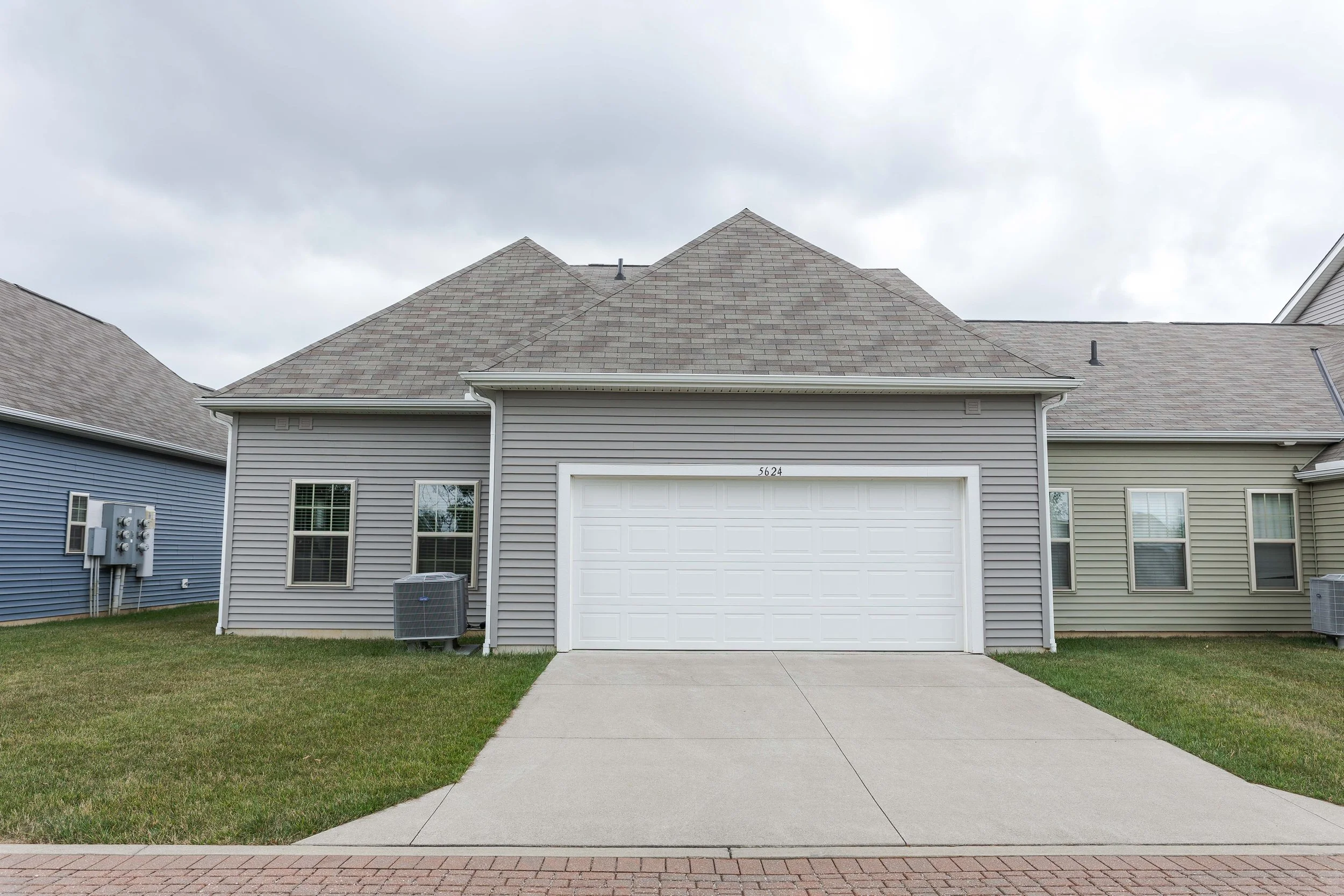 Front view of a modern house with gray vinyl siding, a white garage door, two small windows on the left, and three windows on the right, a sloped roof, and a small lawn on both sides of the concrete driveway under an overcast sky.