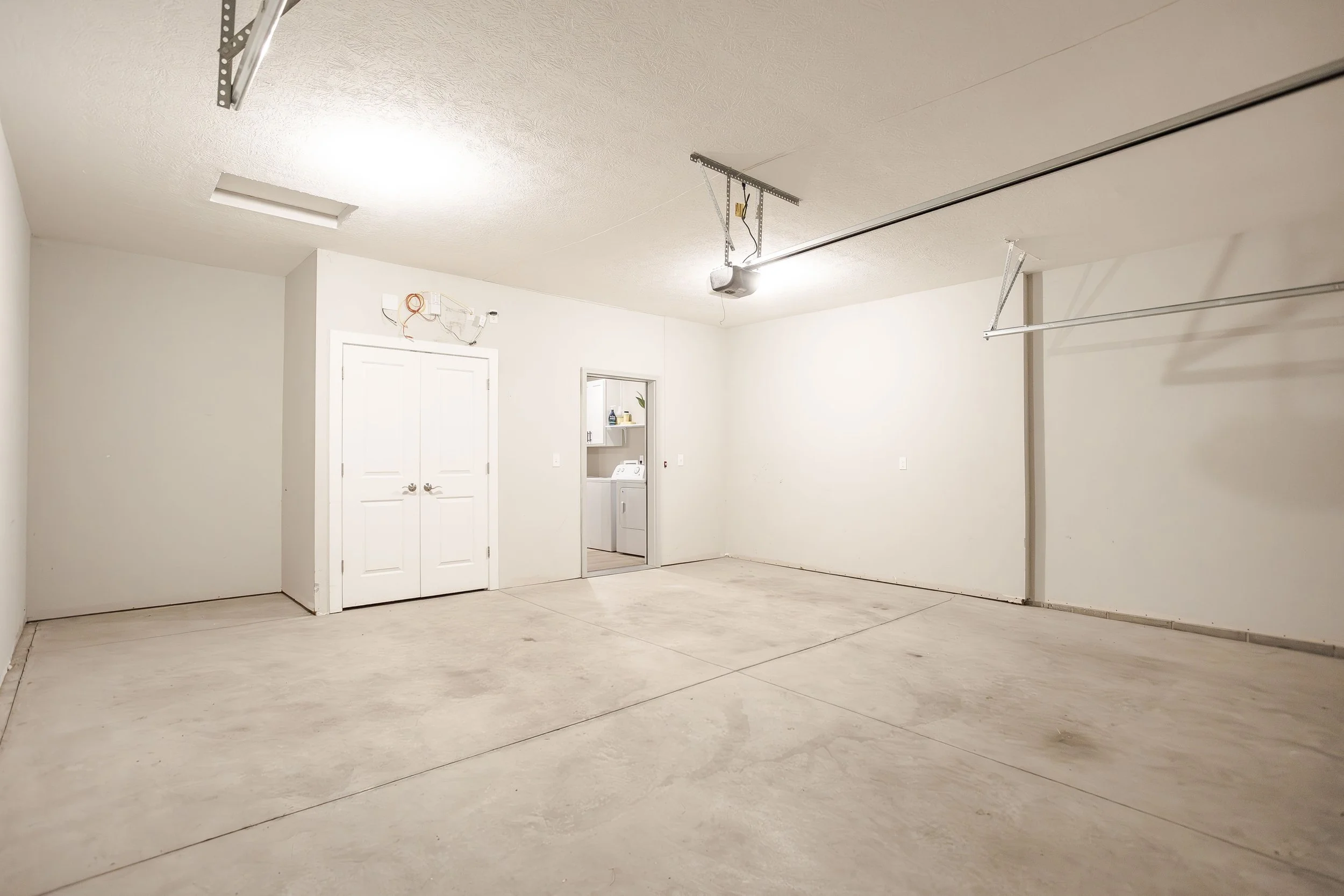 Empty garage with concrete floor, white walls, and ceiling. Two ceiling-mounted garage door openers and tracks are visible. A double closet door and laundry area with washer and shelves are present on the interior wall.