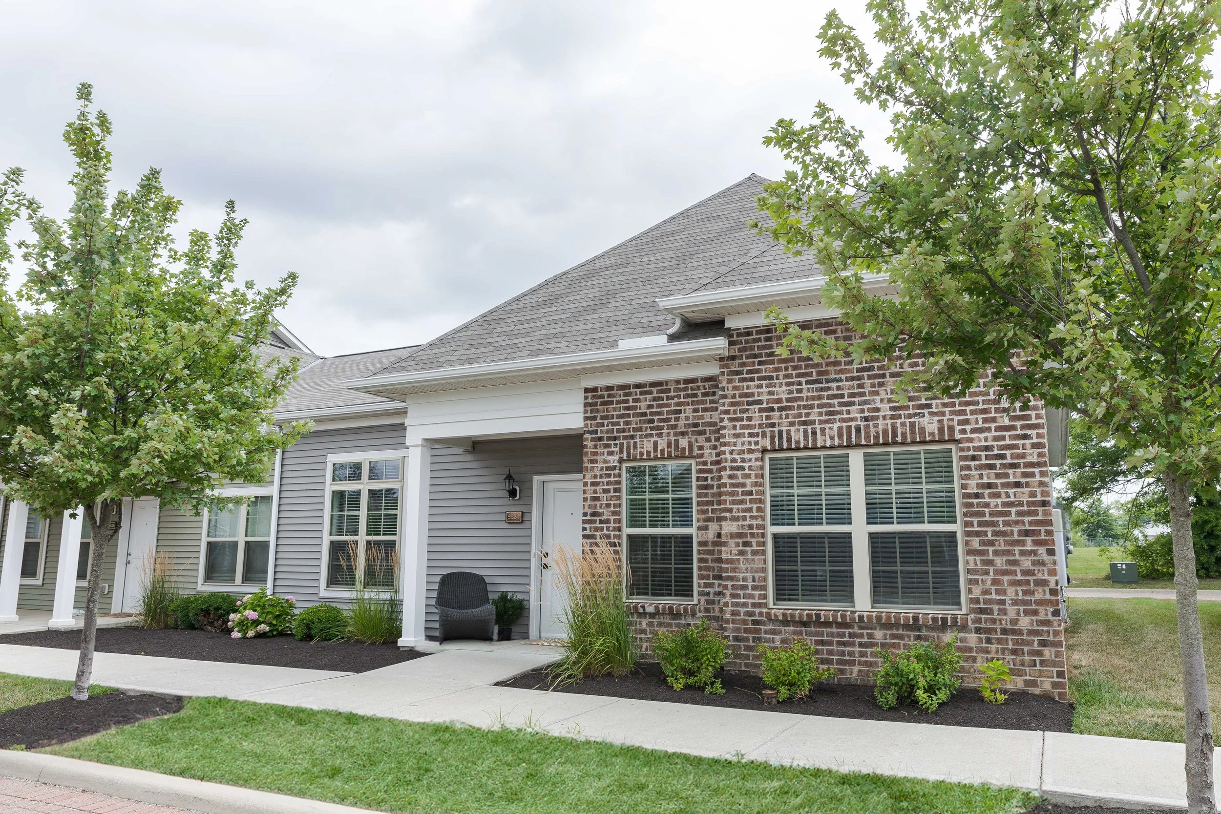 front view of a suburban house with brick and siding exterior, two large windows, a small porch with black chair, manicured lawn, and trees, under cloudy sky.