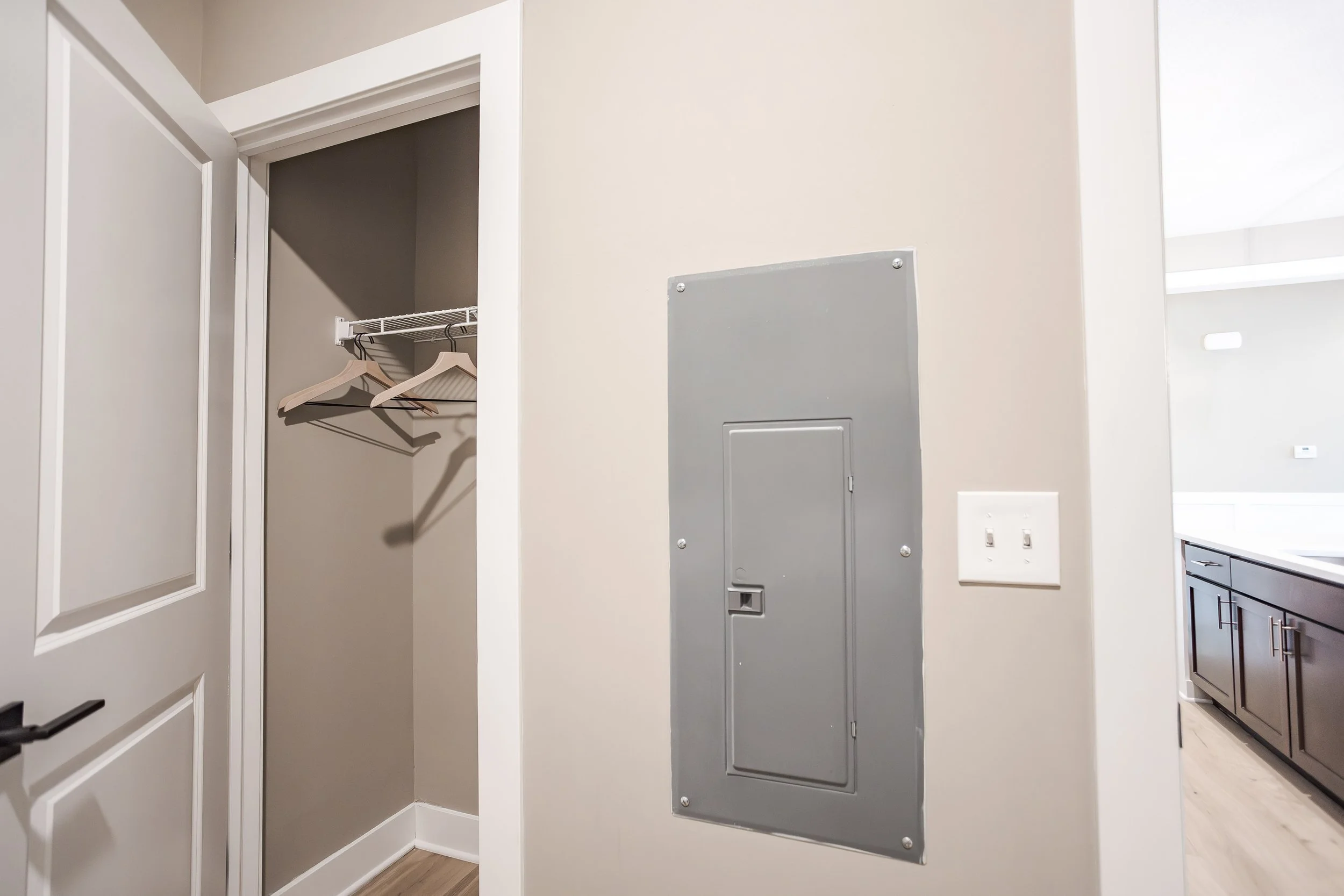 Empty closet with a wire shelf and two wooden hangers, adjacent to electrical panel and light switches in a modern kitchen.