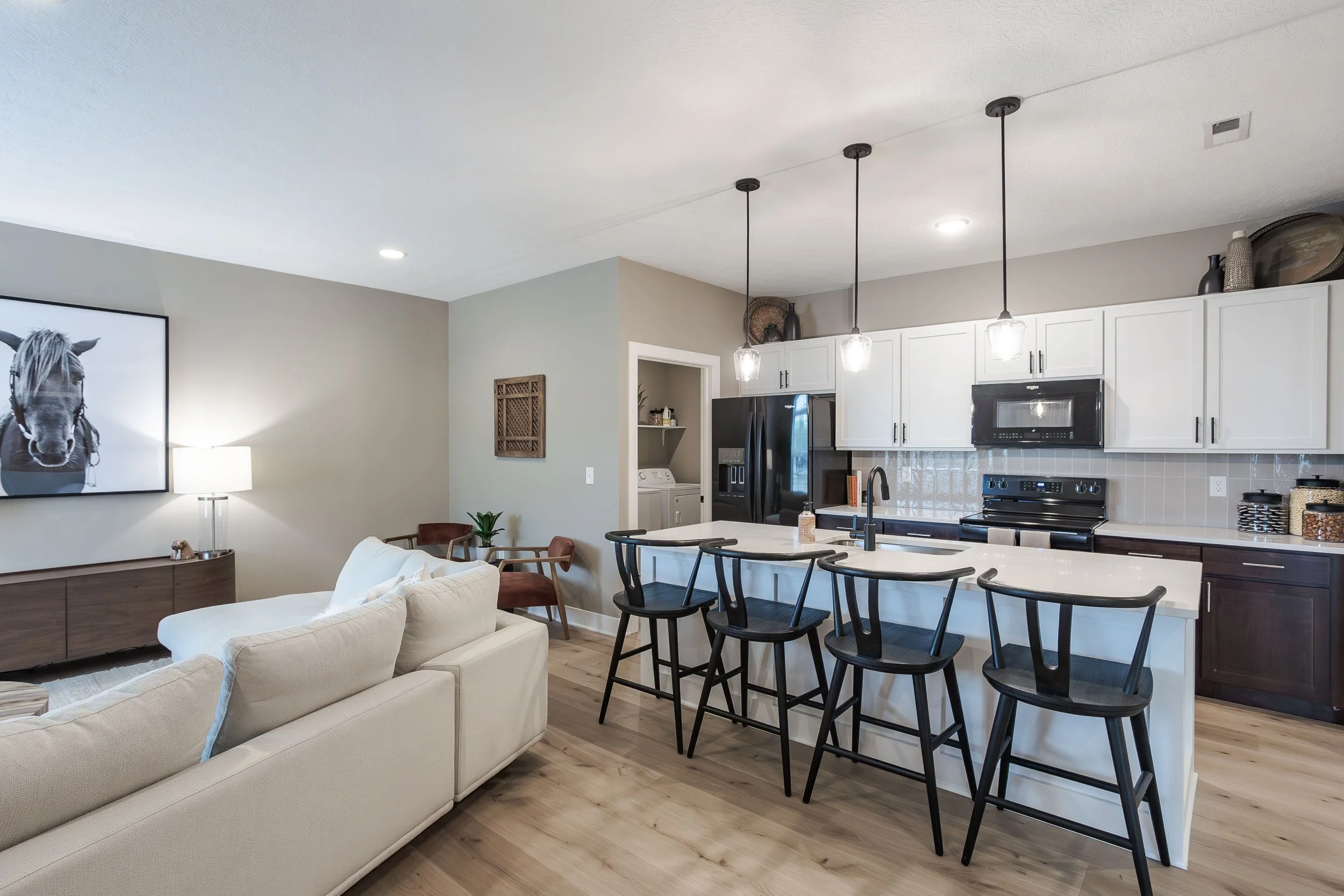 Open-concept living room and kitchen with white cabinets, black appliances, black bar stools, a beige sofa, hardwood floors, and modern lighting fixtures.