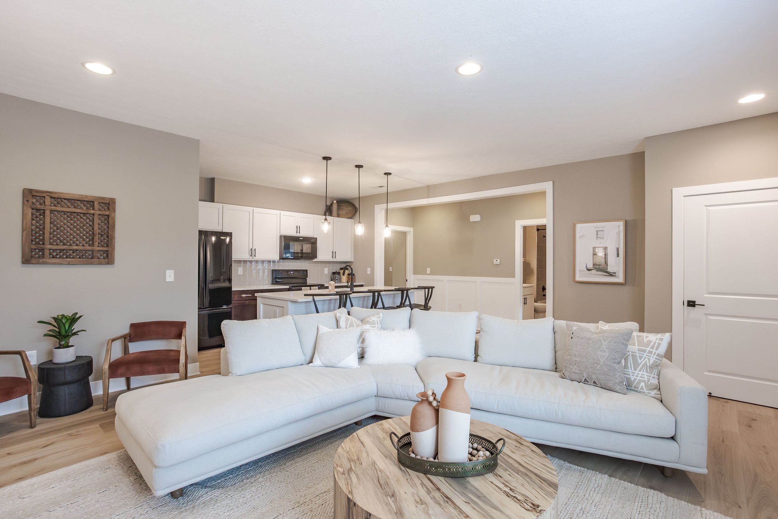 Living room with white L-shaped sofa, wooden coffee table, and neutral decor, open to kitchen with white cabinets and black appliances, pendant lights hanging over the kitchen island.