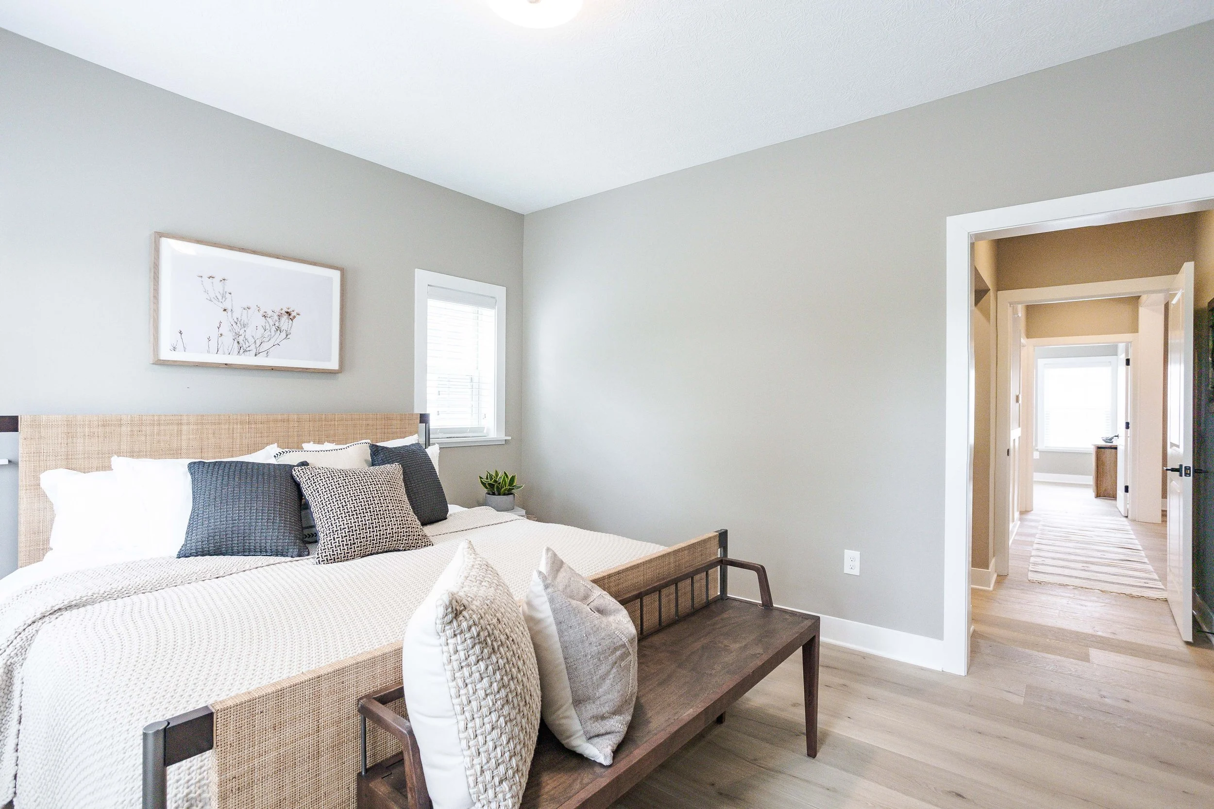 A bedroom with light gray walls, a bed with a rattan headboard, white bedding, and a variety of gray and patterned pillows. A small plant is on the bedside table. There is a window with white blinds, and a framed botanical art piece hangs above the b