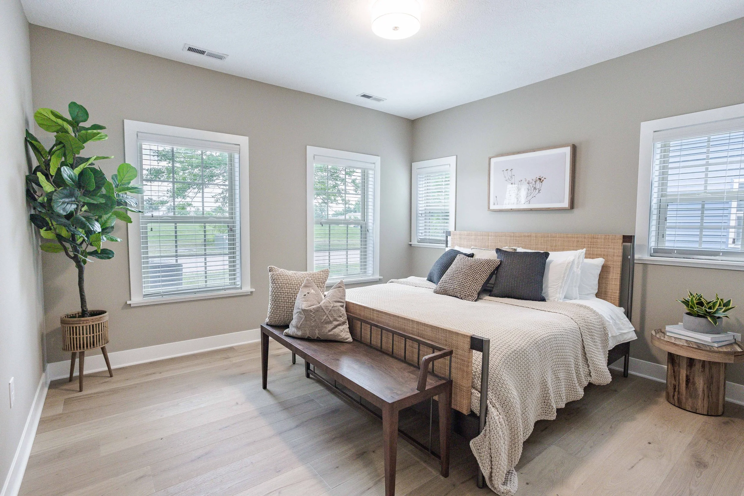 A bright bedroom with five windows, a neatly made bed with beige and dark pillows, a wooden bench at the foot, a side table with a plant, and a large potted plant in the corner.