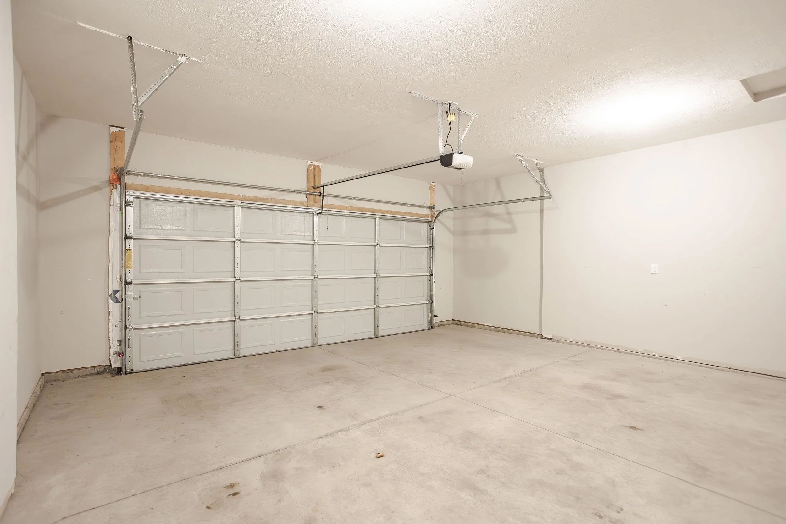 Empty residential garage with white walls, concrete floor, and a closed white sectional garage door, with garage door opener and tracks installed.