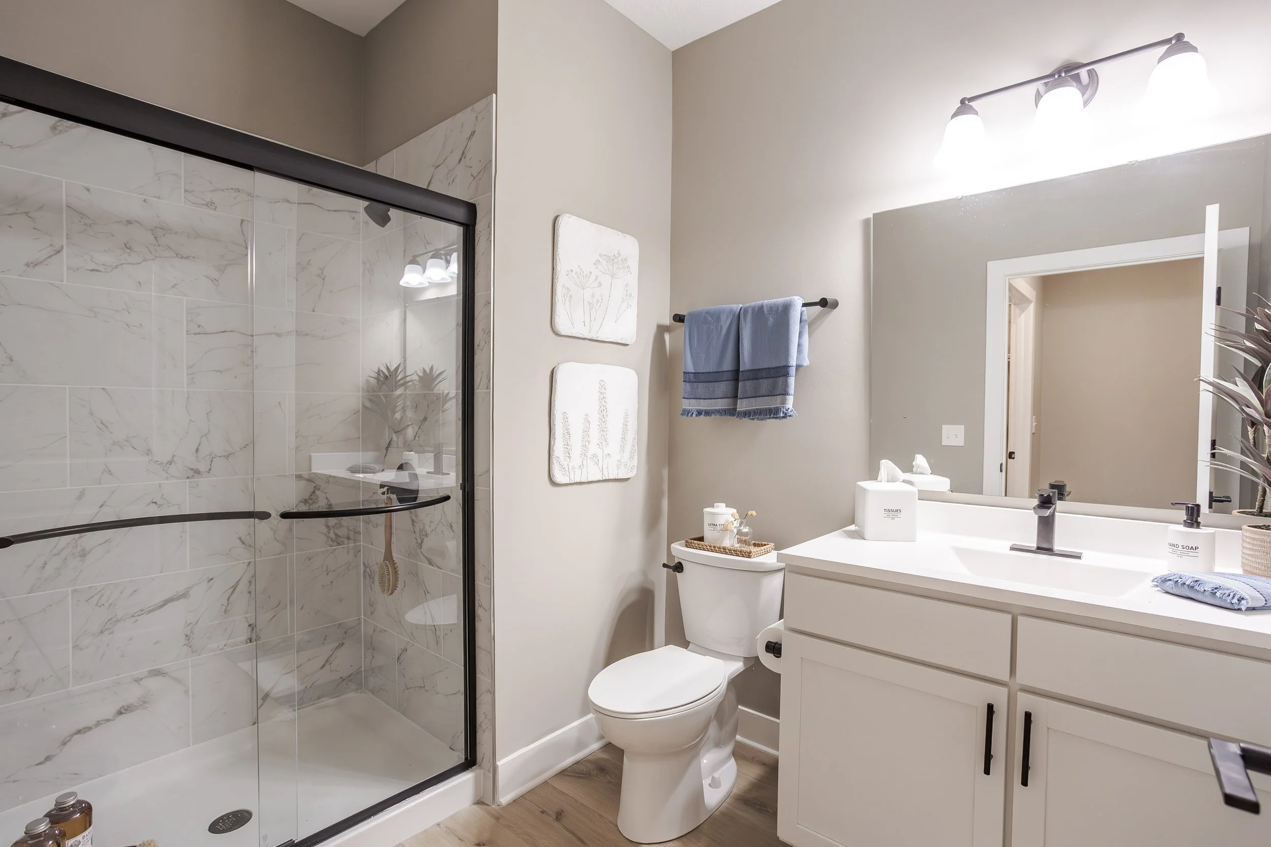 Modern bathroom with a walk-in shower with marble tiles, a white toilet, a white vanity with a mirror, and blue towels hanging on a black towel bar. Decor includes wall art, a tissue box, soap dispenser, and indoor plant.