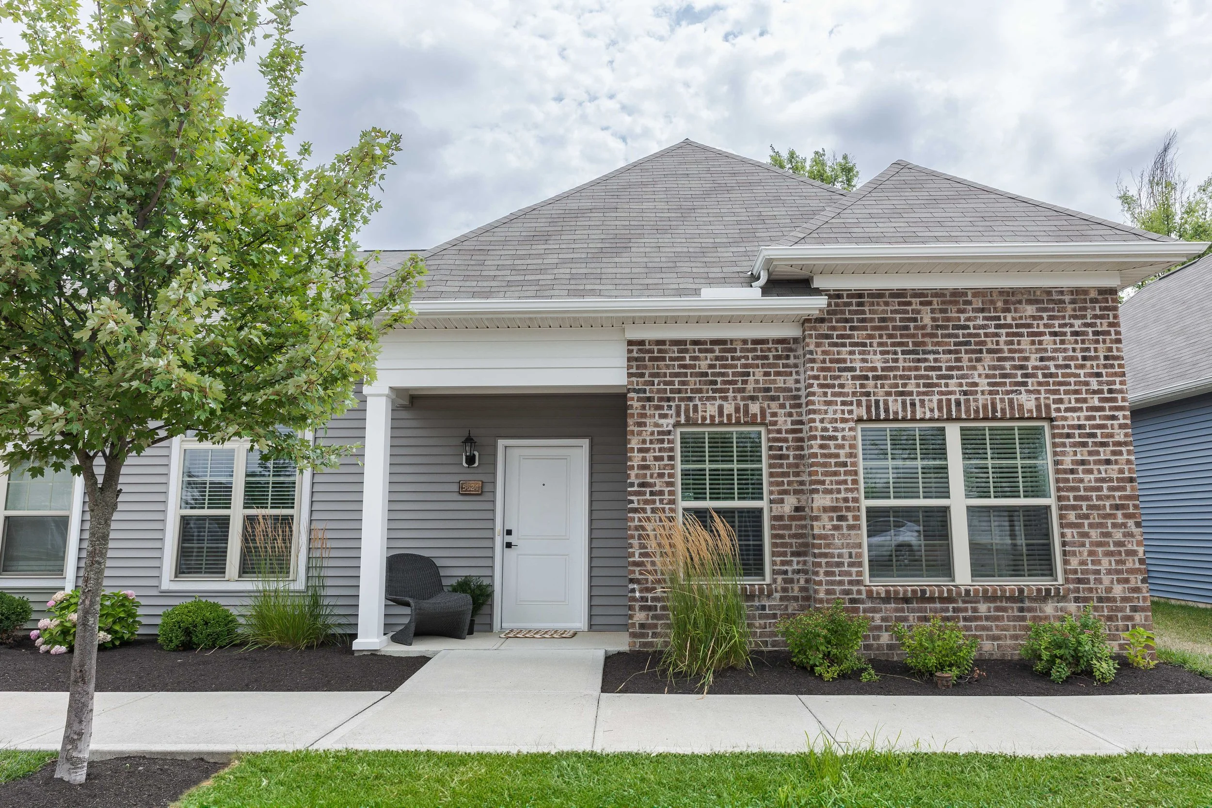 Front view of a house with a brick and siding exterior, gray shingled roof, and a small porch with a black wicker chair and plants, under a partly cloudy sky at The Landon at New Albany Park. 