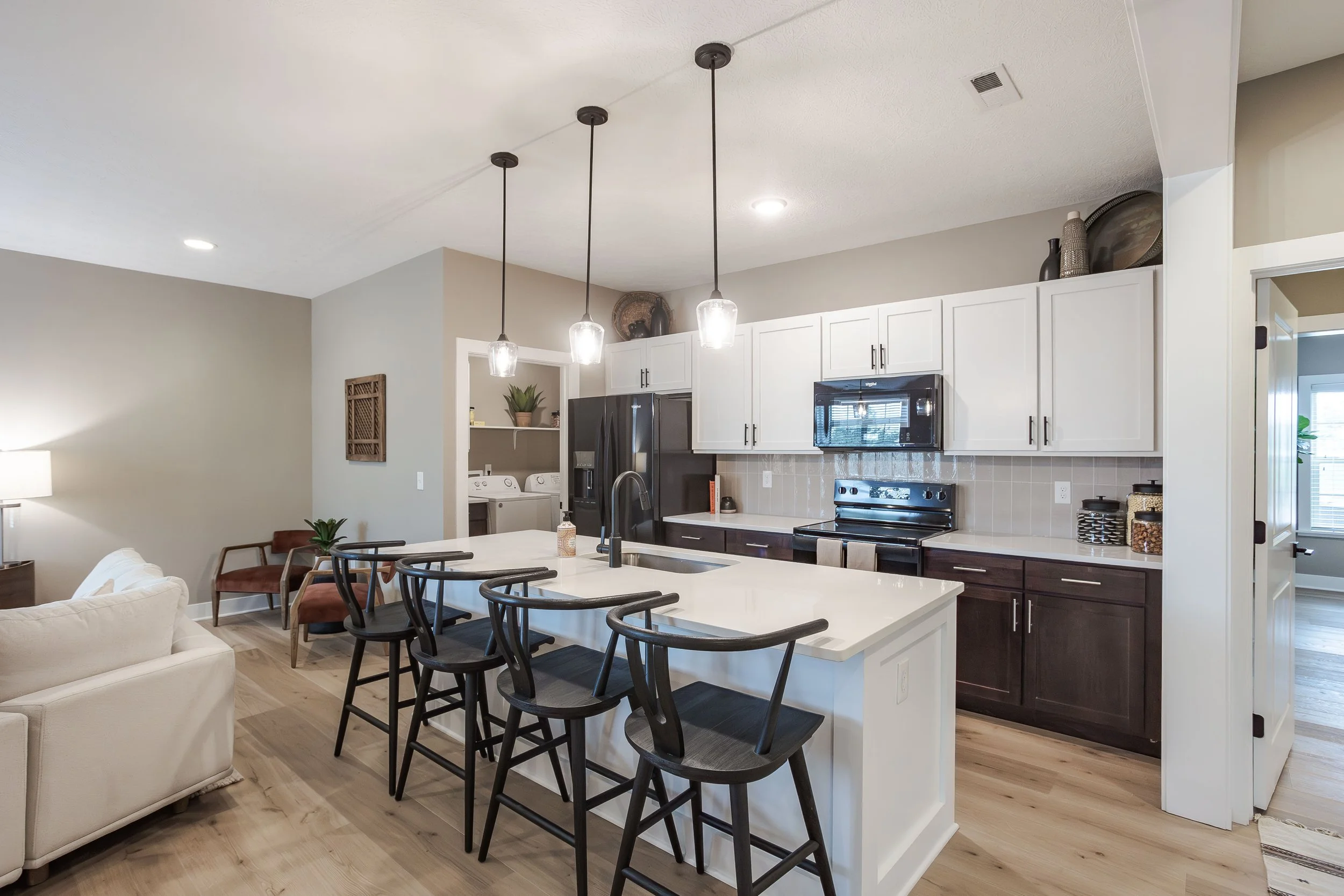 Modern kitchen with white cabinetry, black appliances, a large white island with seating, pendant lights, and a laundry area in the background.