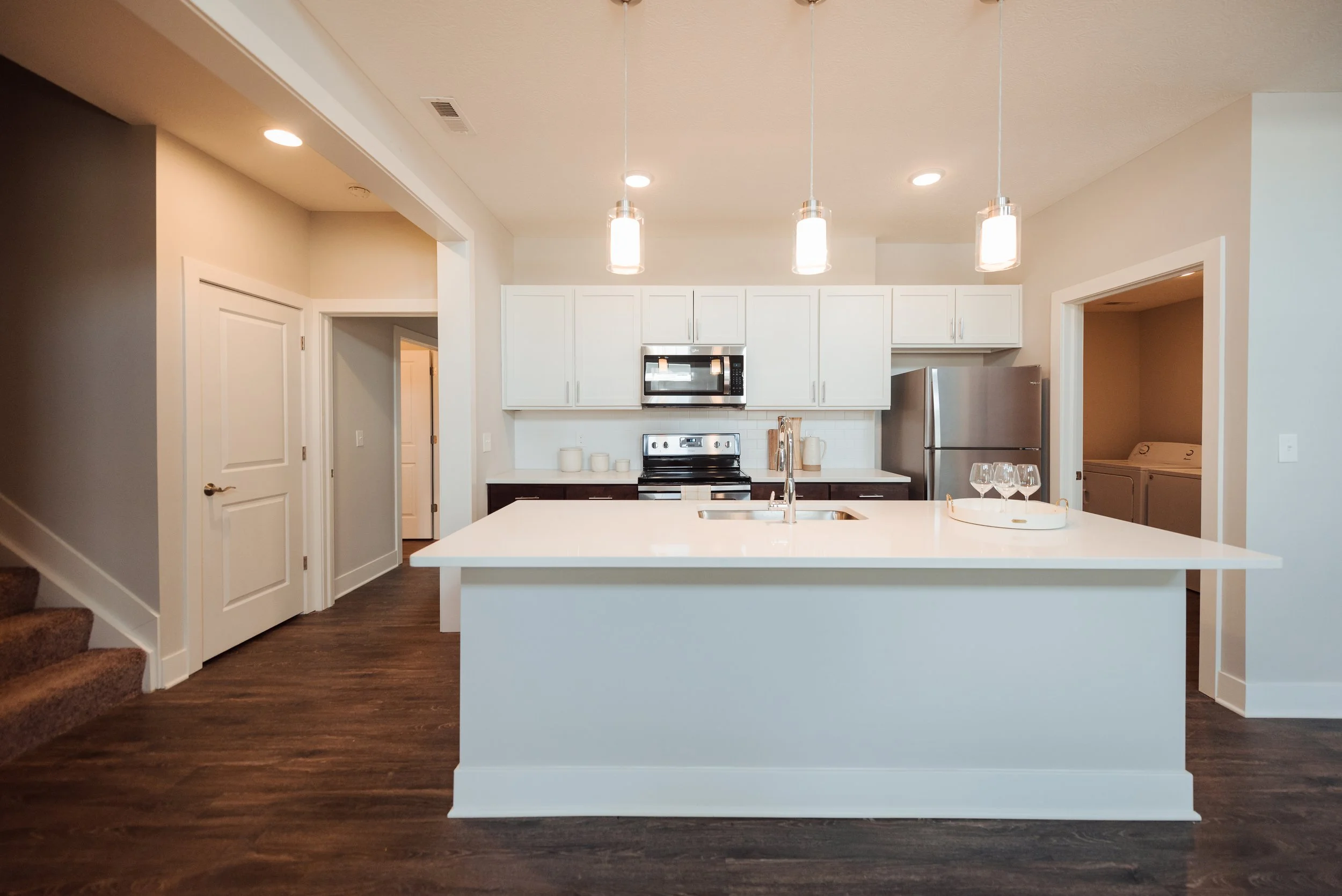 Modern kitchen with white cabinetry, stainless steel appliances, and a large white island with a sink. There are three pendant lights hanging above the island, and a tray with four wine glasses on it. The kitchen has dark wood flooring and an adjacen
