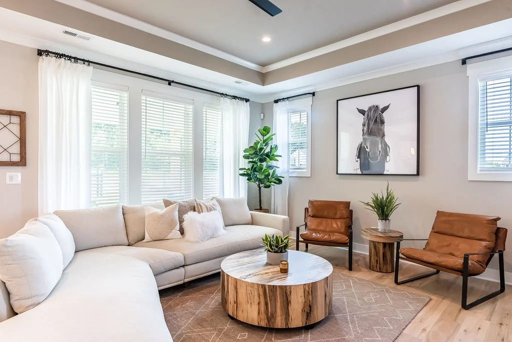 Living room with white sectional sofa, leather armchairs, round wooden coffee table, potted plants, large framed horse photo, white curtains, multiple windows, neutral colored walls, light wood flooring, ceiling fan.