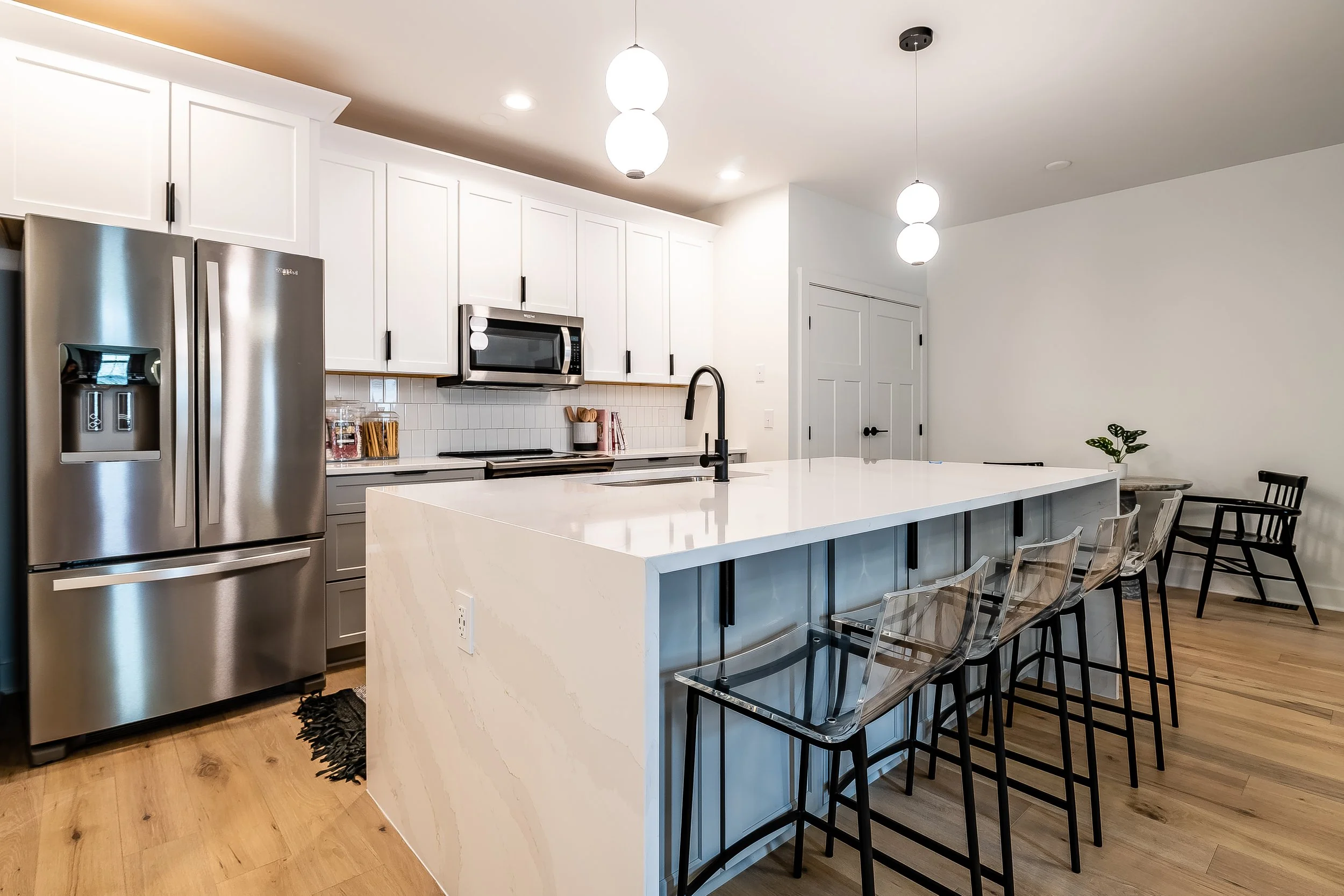 Modern kitchen with white cabinets, stainless steel refrigerator and microwave, black fixtures, a large white island with four transparent bar stools, and a small potted plant in the corner.