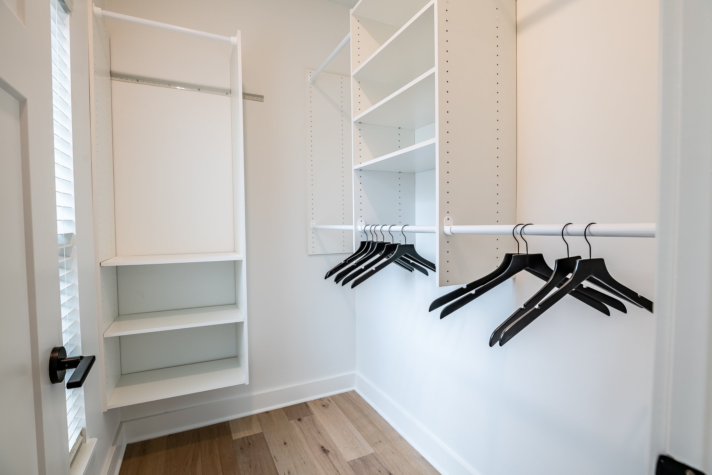 Empty closet with white shelves and hanging rods, black hangers, and hardwood floor.