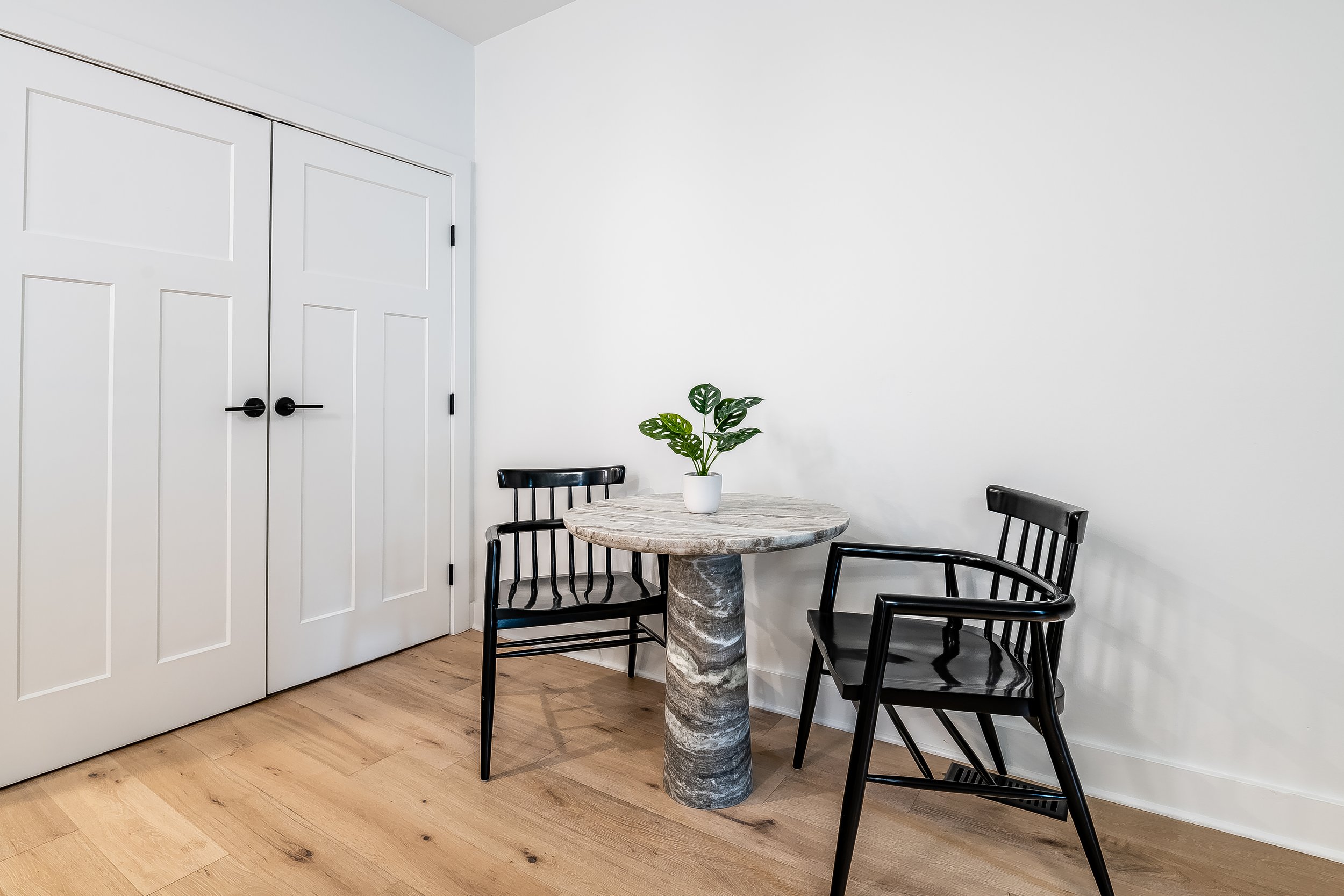 A small round marble table with a potted plant on top, flanked by two black chairs with wooden slats, in a room with a white wall, white closet doors, and a wooden floor.