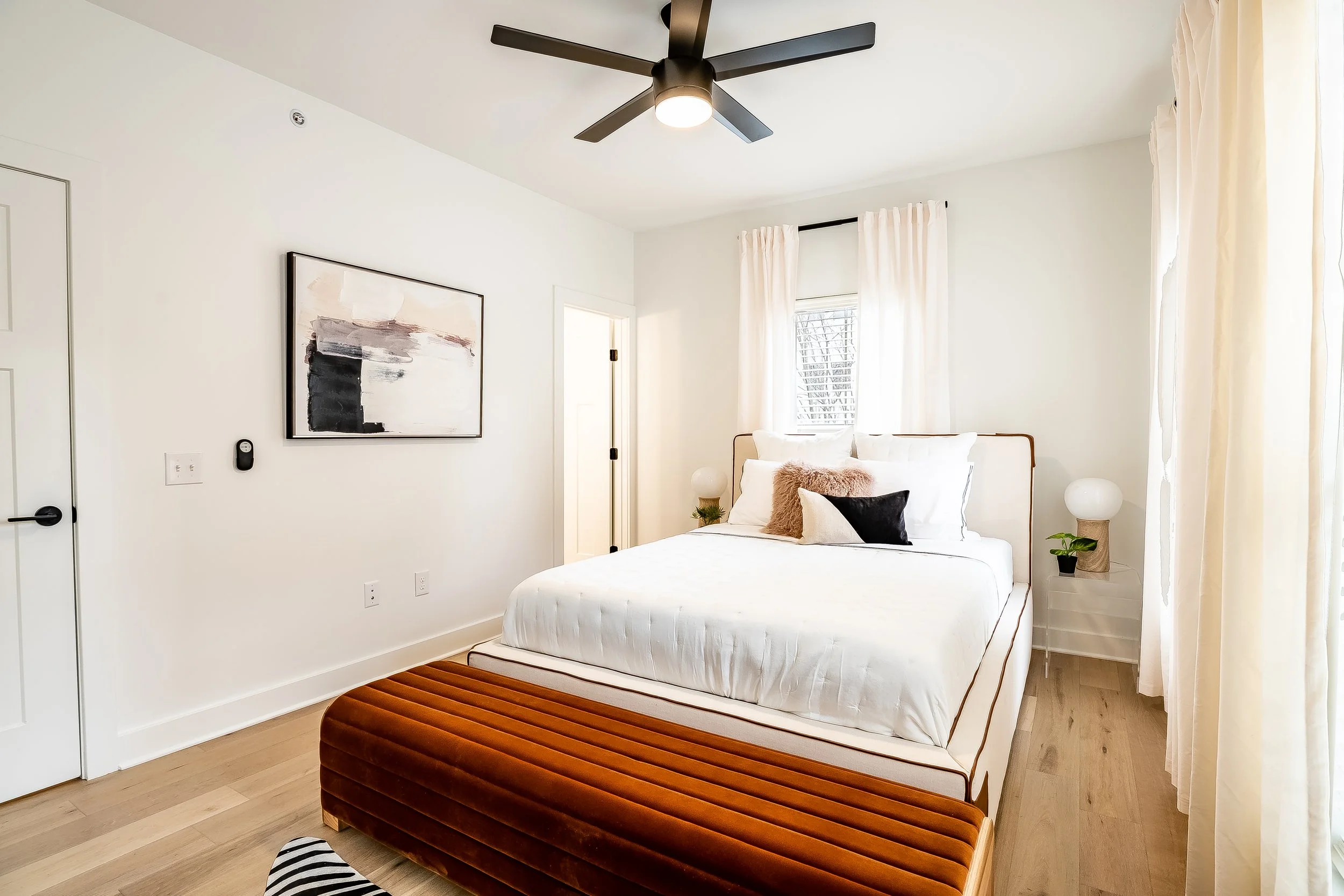 A modern bedroom with a white bed, white curtains, and a brown velvet bench at the foot of the bed. There is a black ceiling fan with a light, and abstract artwork on the wall.