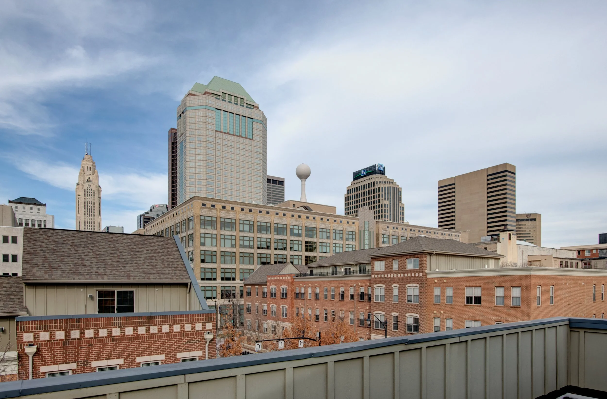City skyline with tall modern buildings, including a beige brick building, and a gray rooftop railing in the foreground under a cloudy sky.