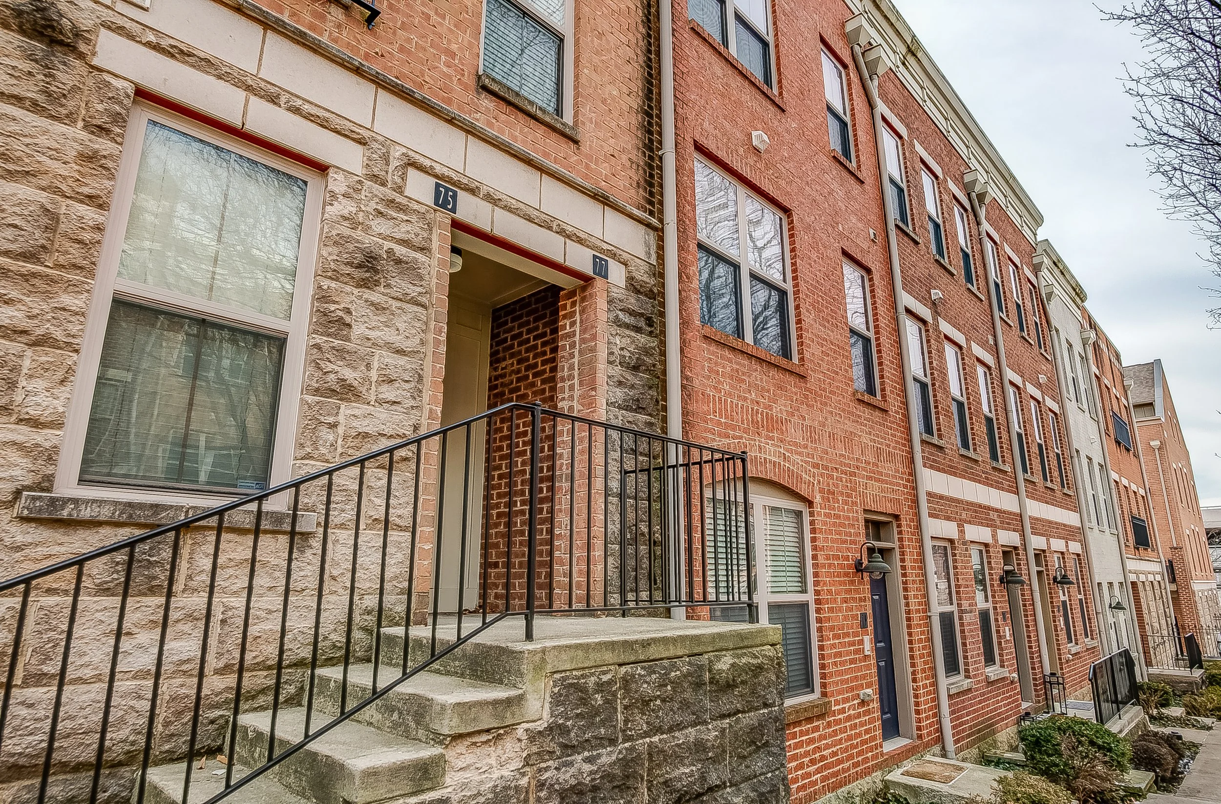 Row of brick and stone apartment buildings with stairs and black railings, front doors, and multiple windows, under an overcast sky.