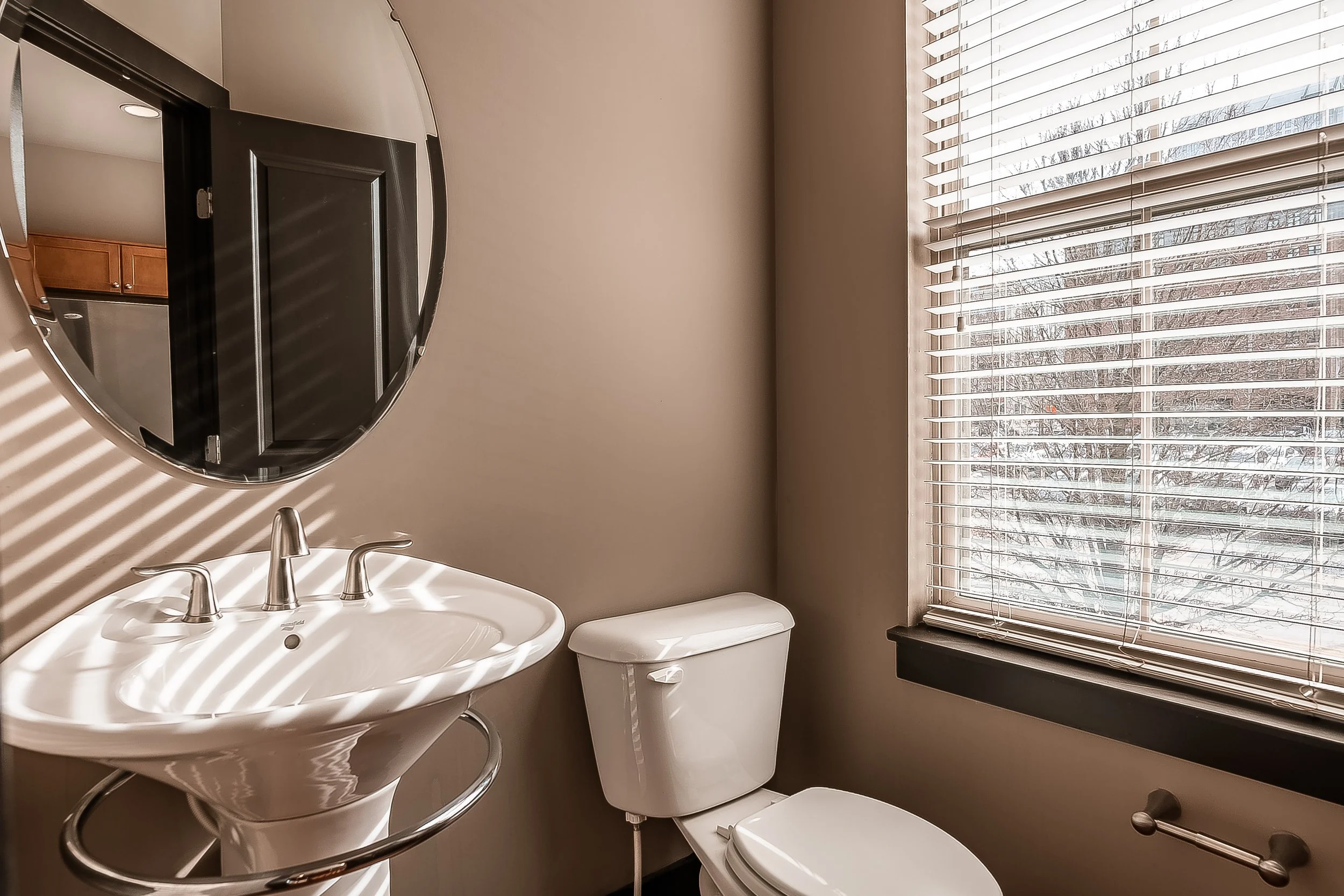 A small bathroom with a pedestal sink, an oval mirror above it, a toilet next to a window with blinds, and a towel bar below the window. The window shows a snowy outdoor scene.