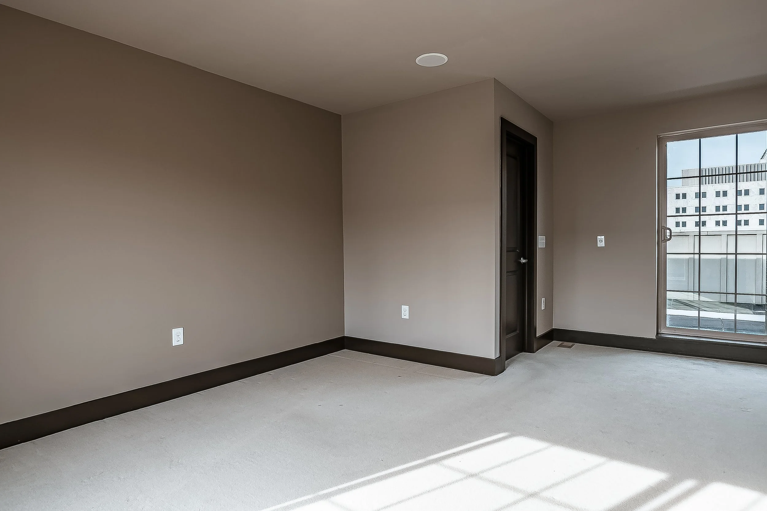 Empty room with beige walls, dark trim, large window with city view, and light-colored carpet.