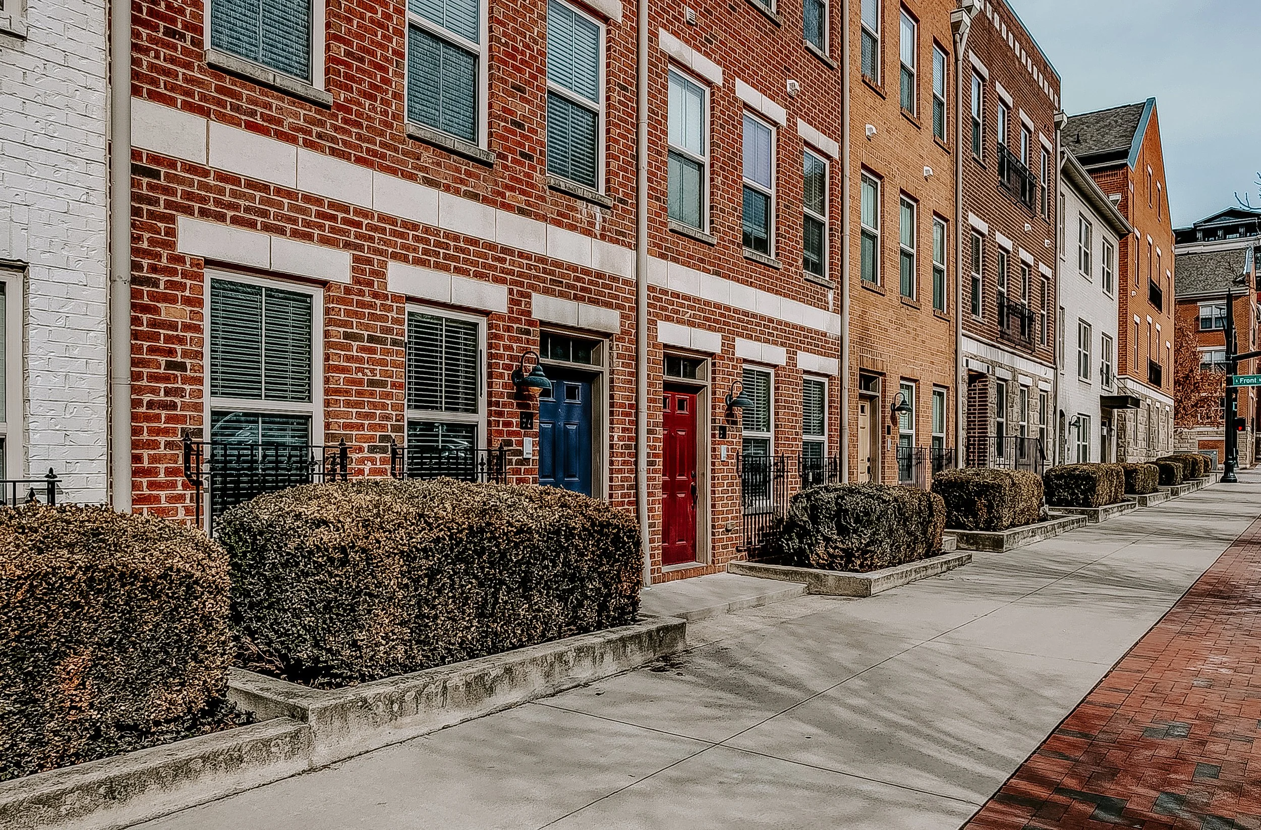 A row of modern brick townhouses with colorful doors and well-kept bushes along the sidewalk.