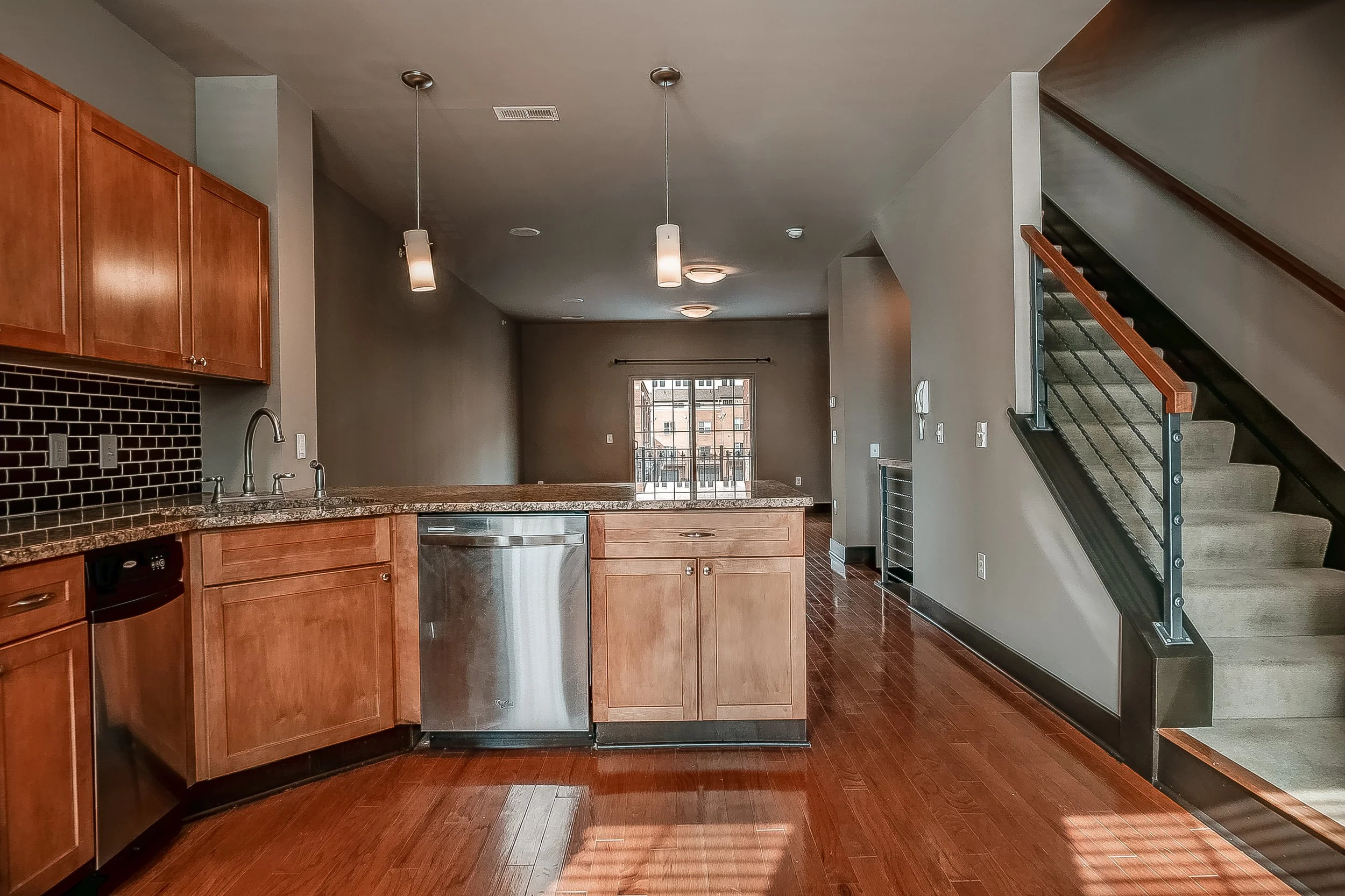 Empty kitchen and living area with hardwood floors, wooden cabinets, granite countertops, black tiled backsplash, stainless steel dishwasher, staircase with wooden handrail, and sliding glass door leading to balcony.