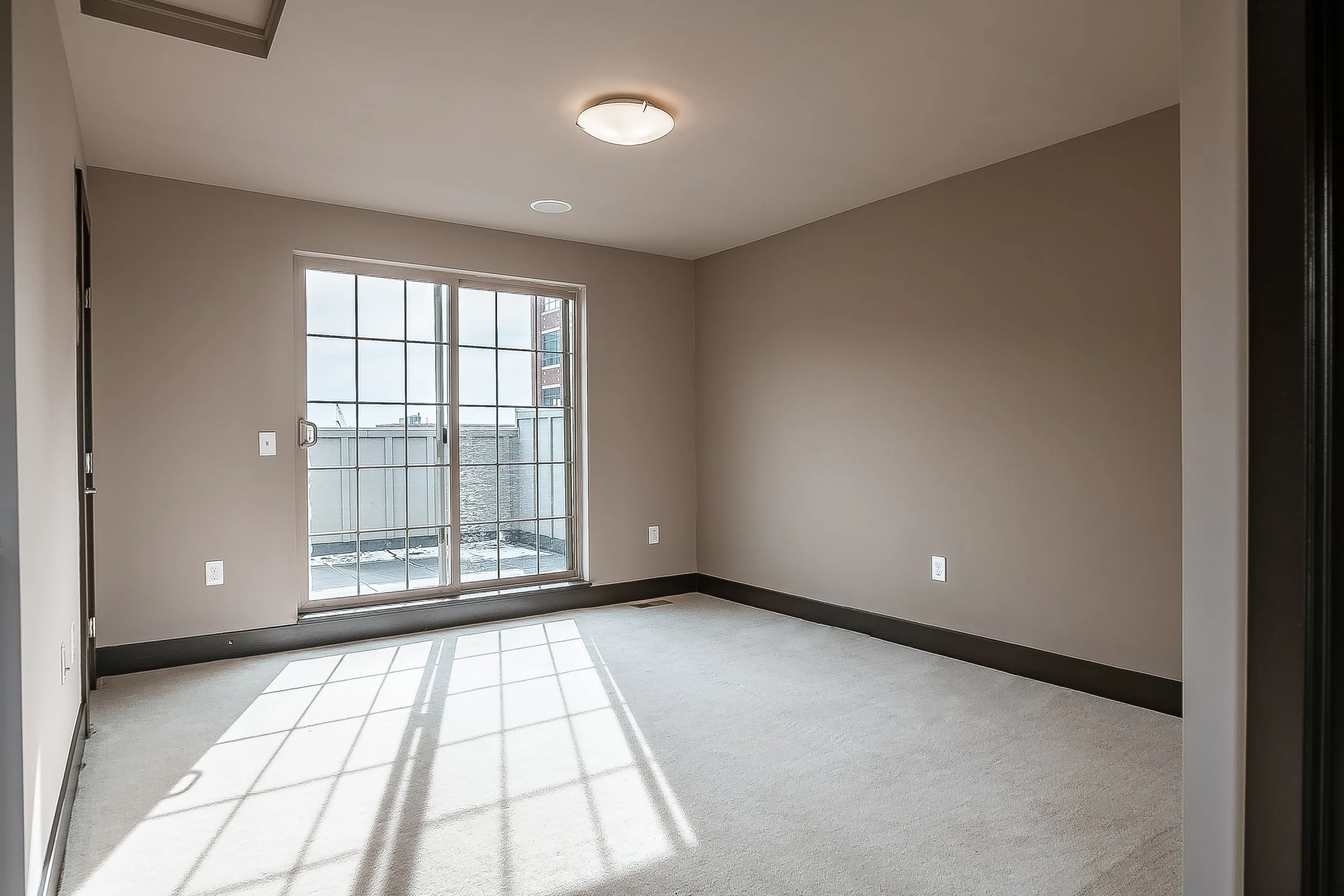 Empty room with beige walls, a sliding glass door leading to a small balcony, and sunlight casting shadows on a light-colored carpet.