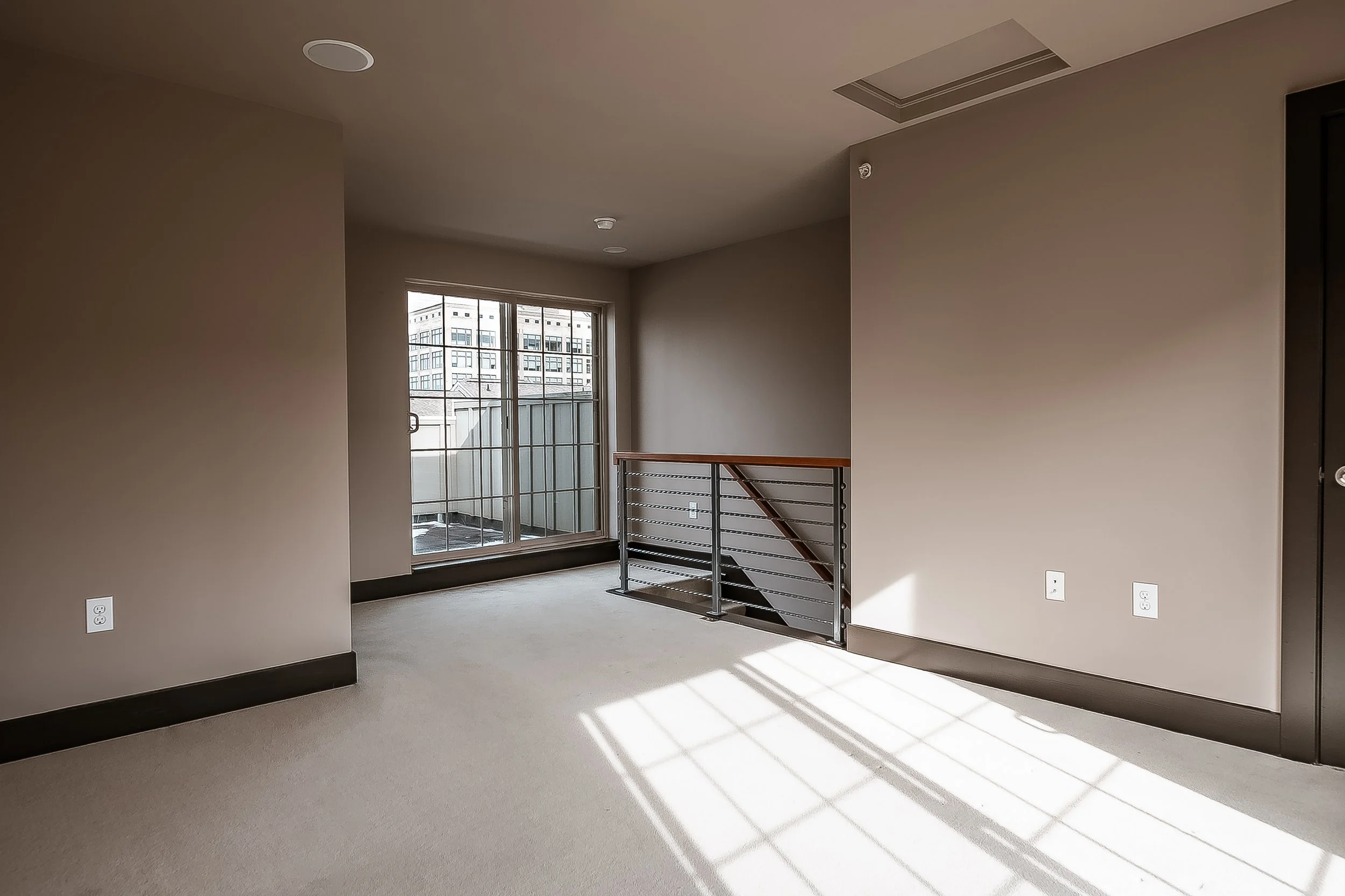 Empty modern apartment room with beige walls, gray carpet, sliding glass door leading to balcony, railing at stairwell, and sunlight casting shadows on floor.