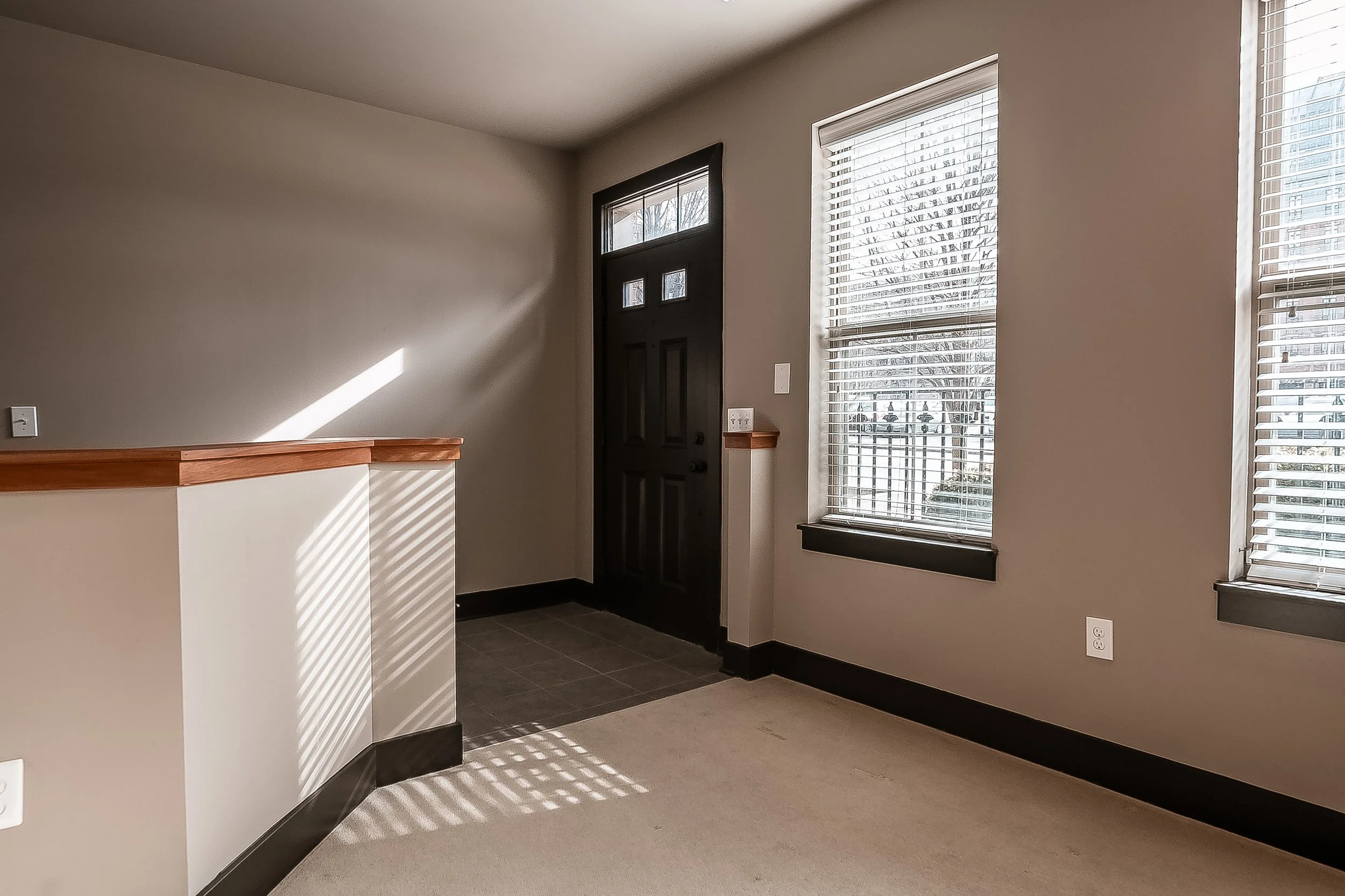 Empty room with beige walls, two large windows with blinds, and a black front door with decorative glass. Carpeted flooring and a small section of tiled flooring near the entrance.