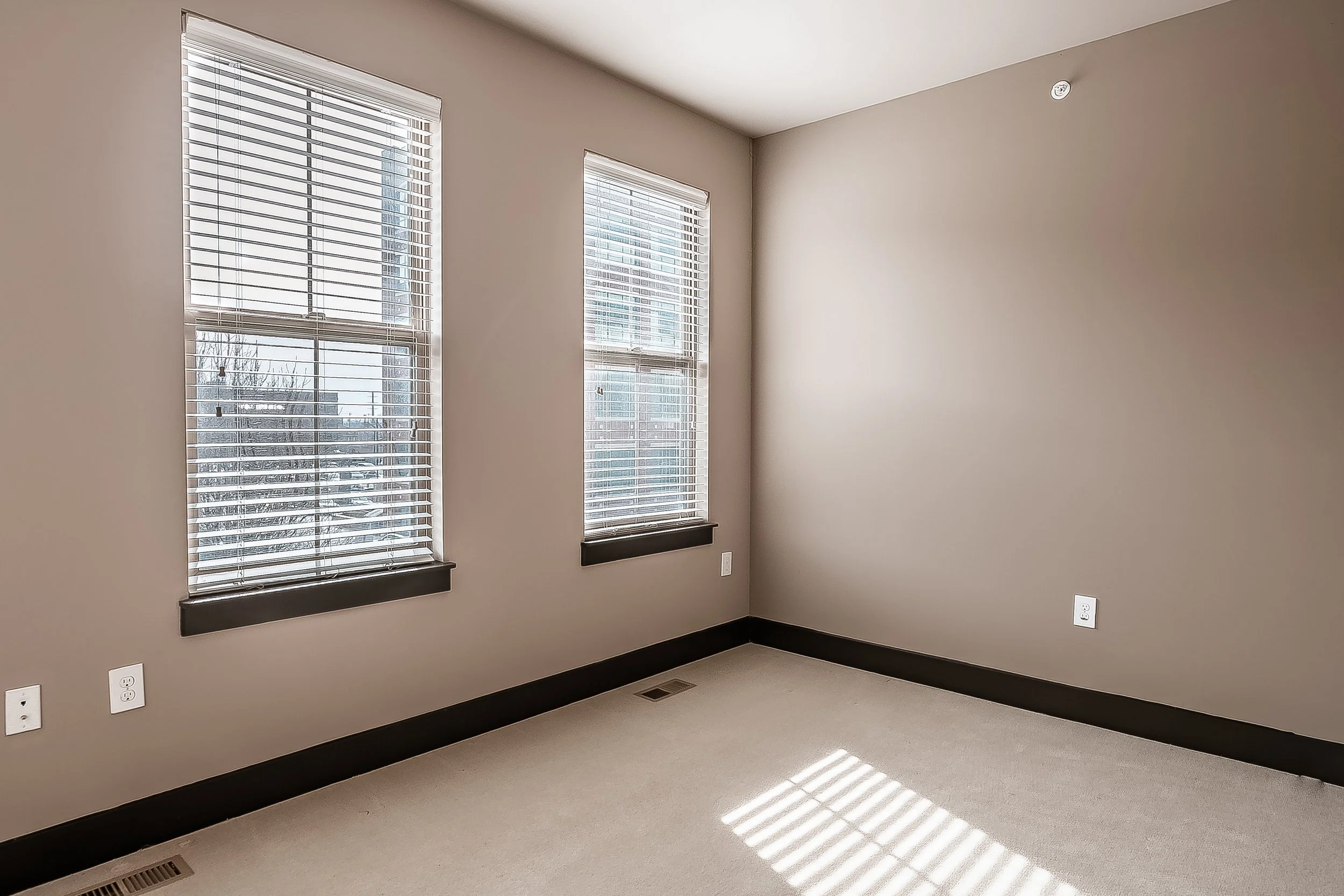 Empty room with beige walls, two large windows with white blinds, black window sills, beige carpet, and electrical outlets.