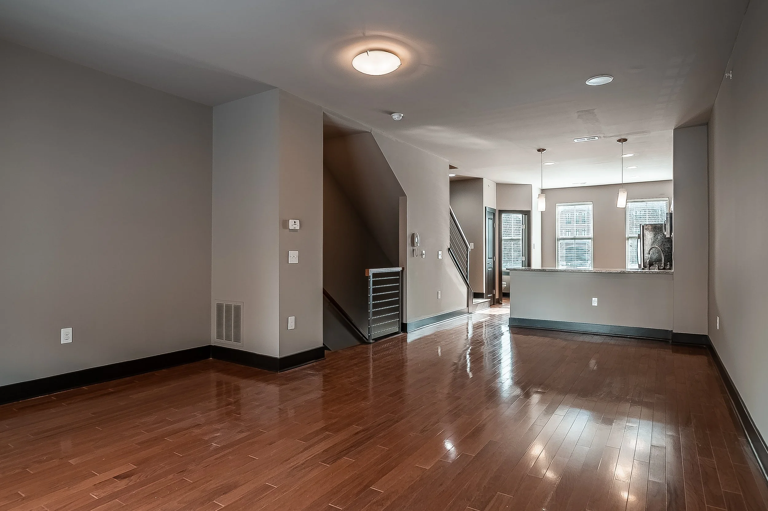Empty living room with hardwood floors, beige walls, and large windows in the background, with an open kitchen area including a granite countertop and pendant lights.