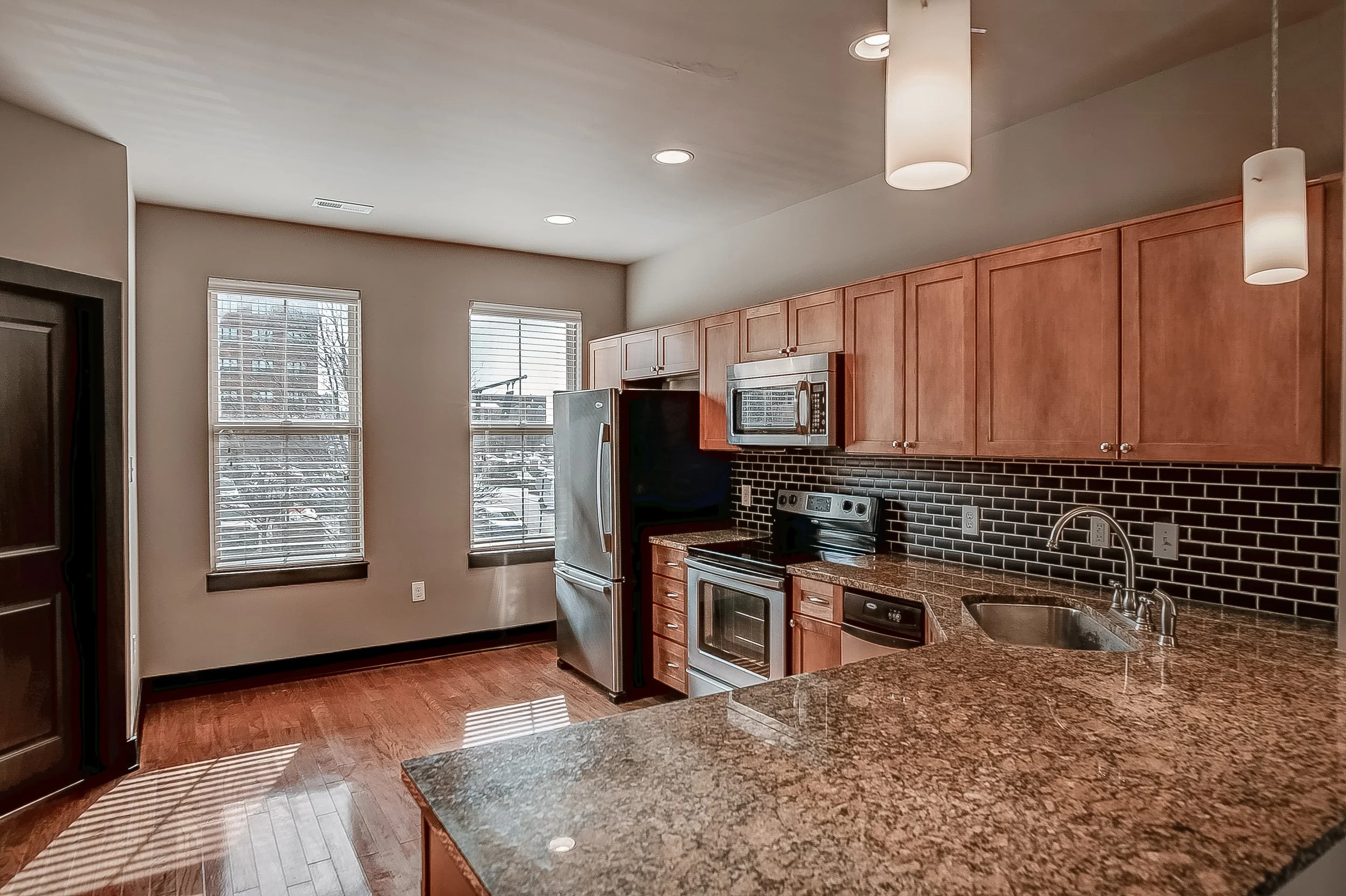 Kitchen with wooden cabinets, black tile backsplash, granite countertops, stainless steel refrigerator, microwave, oven, and dishwasher. Two windows let in natural light, and there are pendant and ceiling lights.