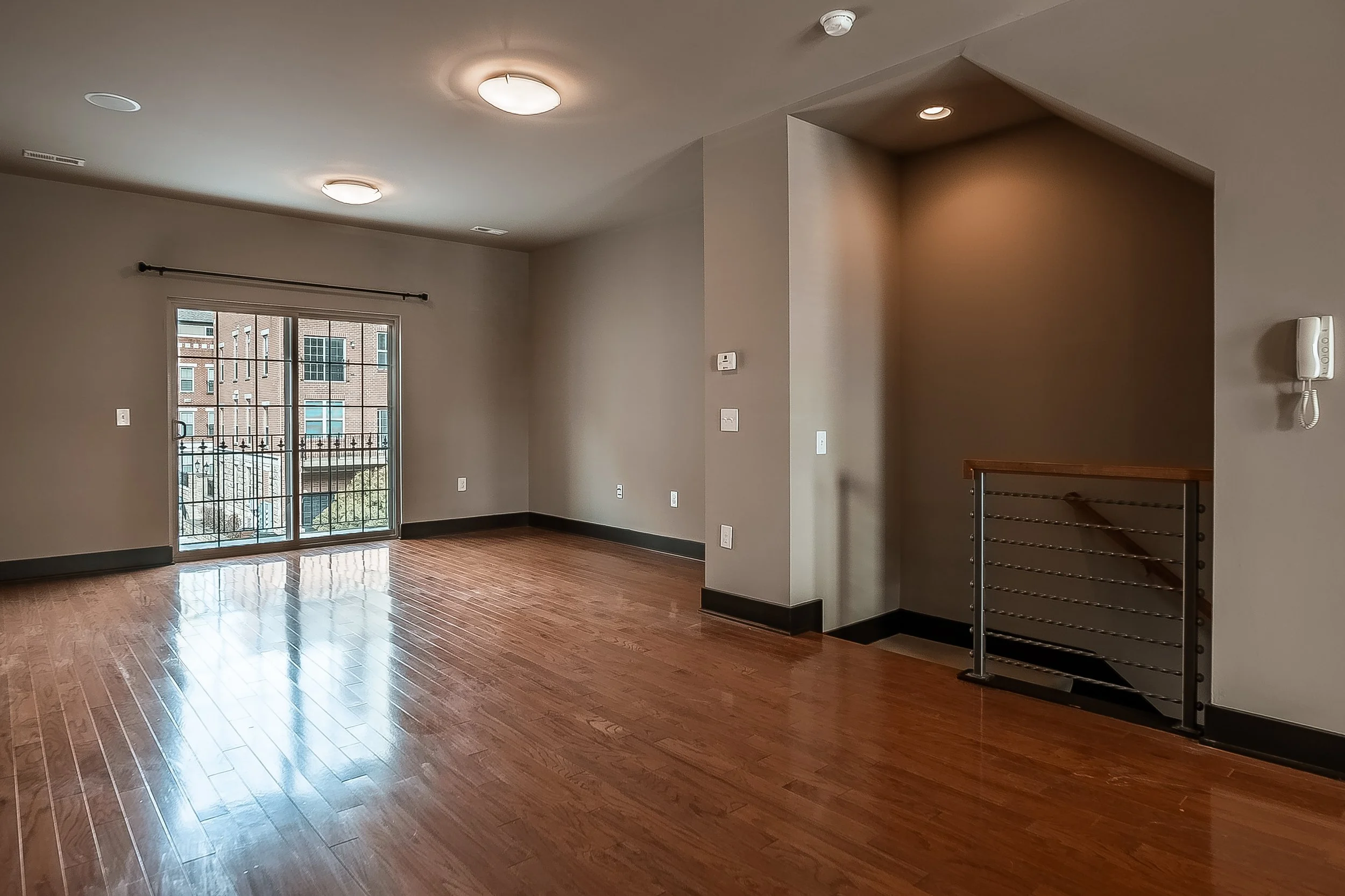 Empty living room with hardwood floors, sliding glass door leading to balcony, gray walls, wall-mounted thermostat, intercom, and stairs with metal railing.