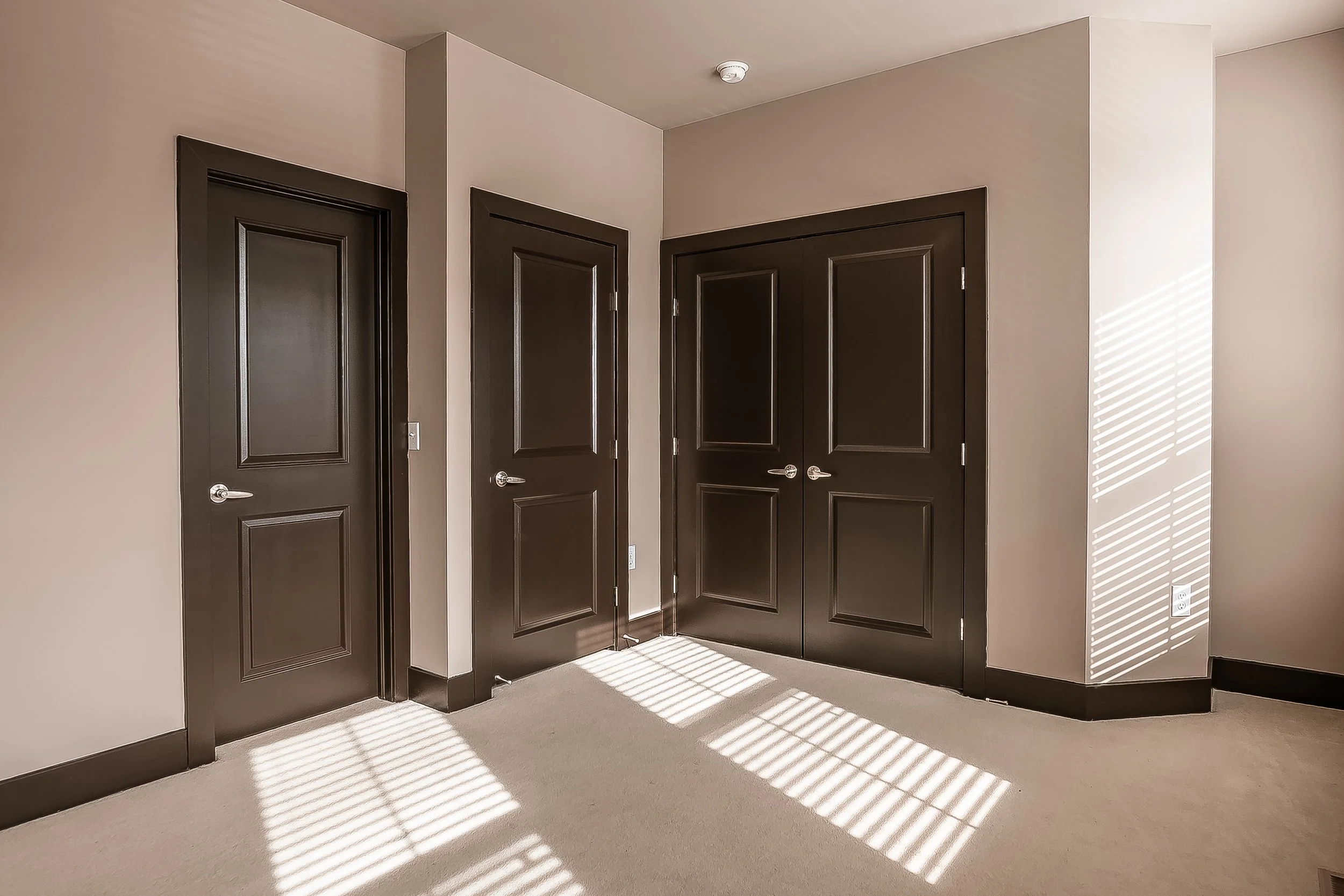 A room with beige walls and carpeted floor, featuring three closed dark brown doors and sunlight creating striped shadows on the walls and floor.