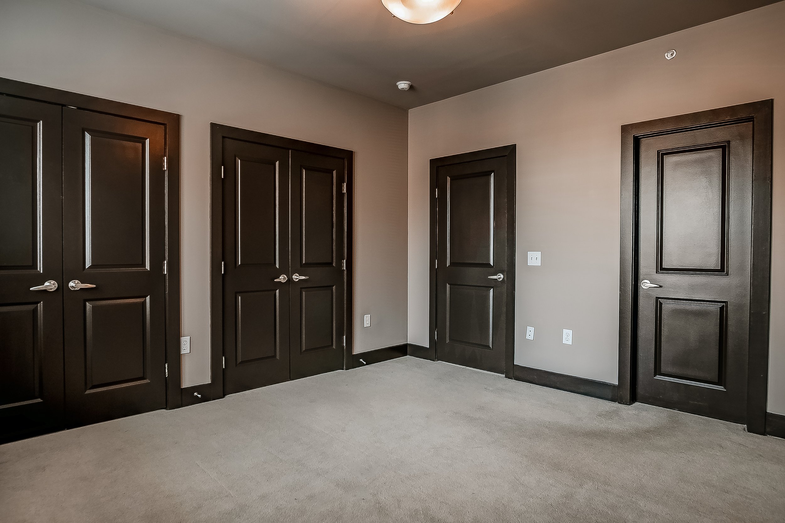 Empty room with beige carpet, light brown walls, and four dark wooden doors.