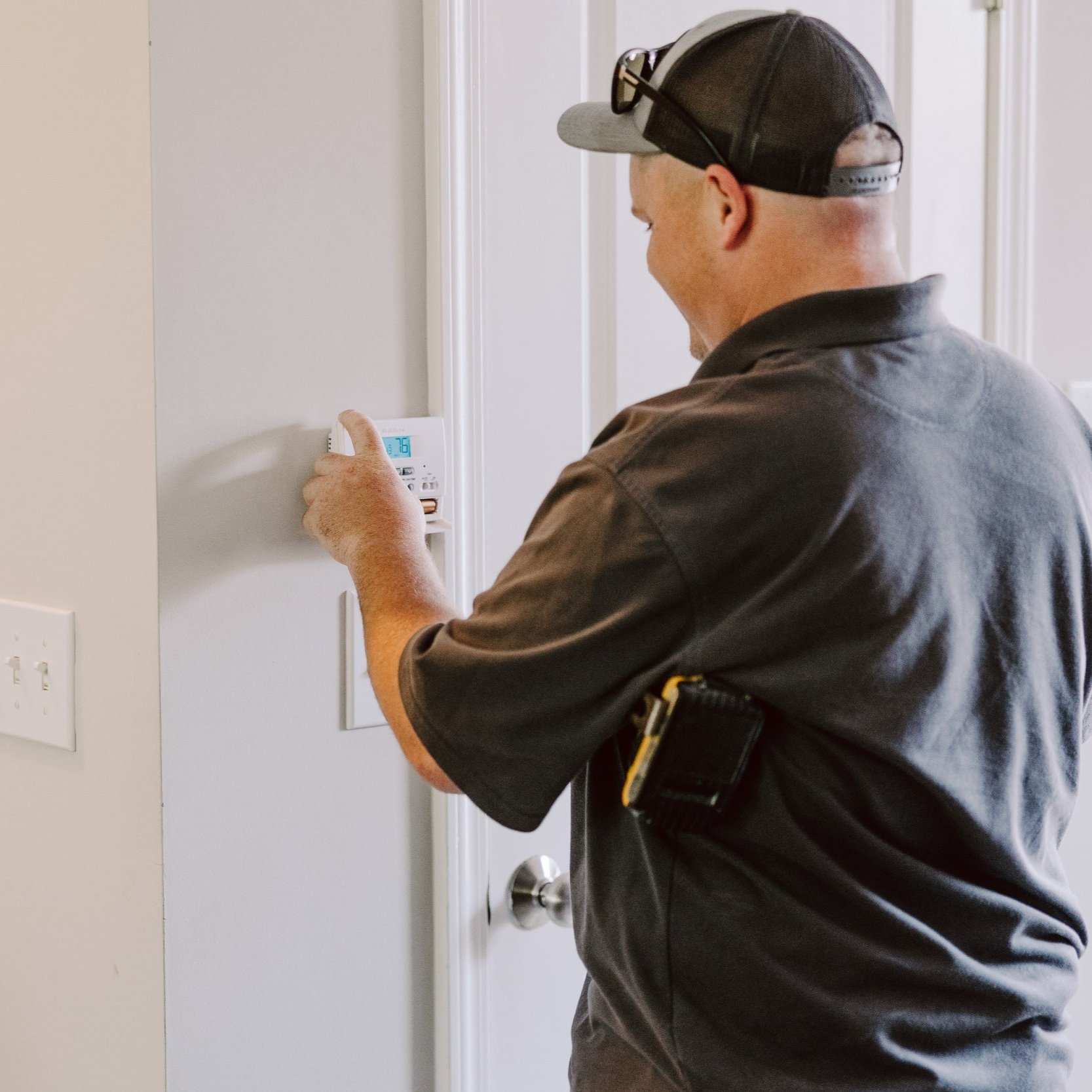 A man in a dark shirt and cap adjusts a thermostat on a wall.